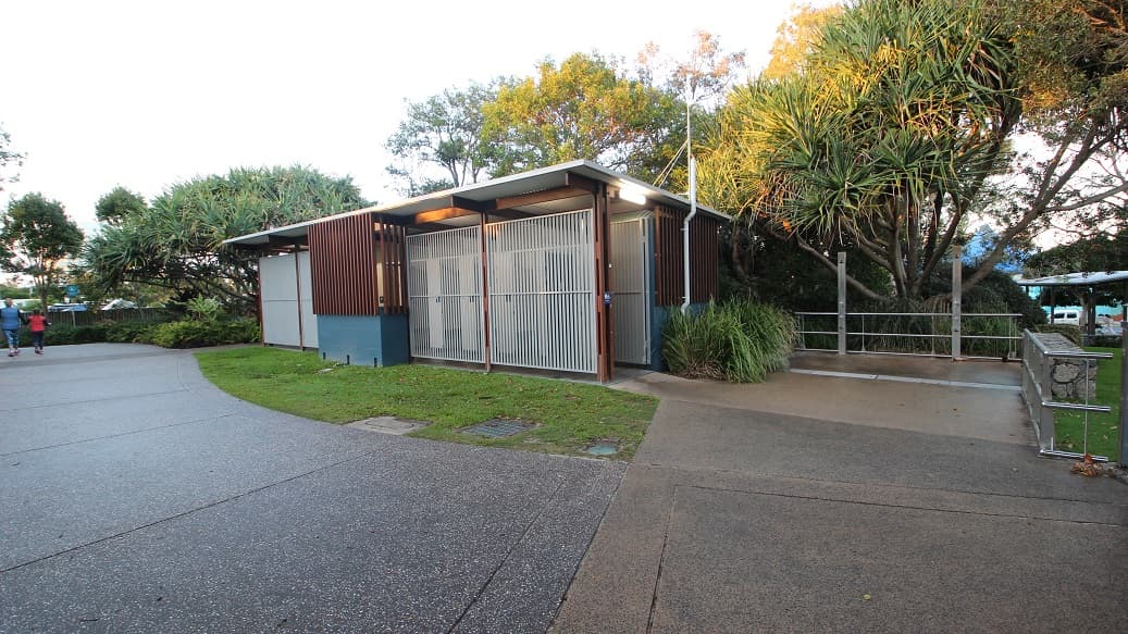 On the left, a paved walkway curves past grass and dense green trees. In the middle, a modern structure with brown slatted panels, white metal doors, and a slightly overhanging roof sits on a green lawn. On the right, a continuation of the building is surrounded by shrubs, a metal railing, and another paved walkway leading slightly downward. The floor consists of a mix of concrete and grass, and the ceiling is the building's roof with a slim guttering system.