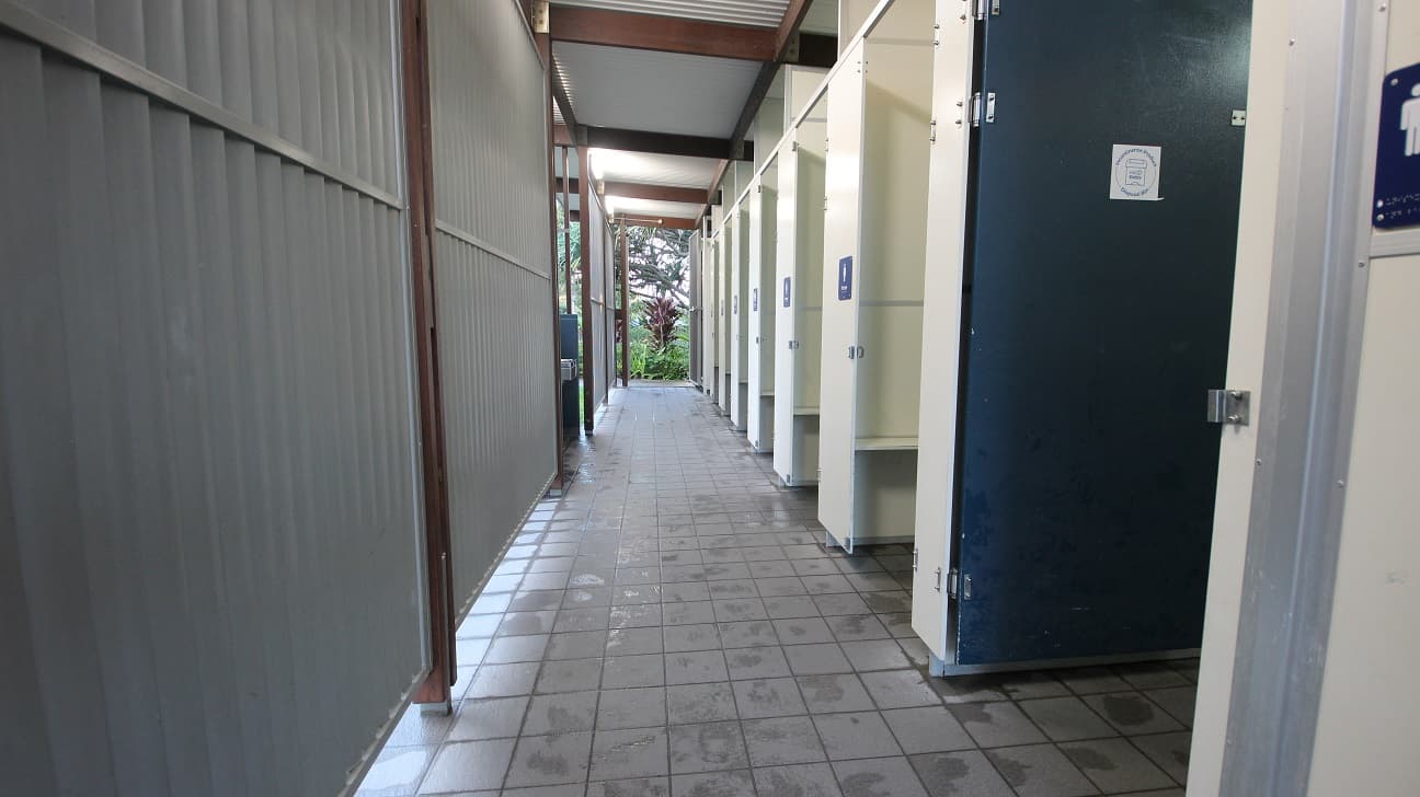 On the left, a grey corrugated metal wall runs the length of the walkway, bordered by wooden beams. On the right, a row of cream-coloured cubicle doors stands partially open, with a dark blue closed door nearest the foreground. In the background, plants and trees are visible through the open end of the walkway. The floor is tiled with square grey tiles, showing damp patches, and the ceiling is a flat, white panelled roof supported by wooden beams.