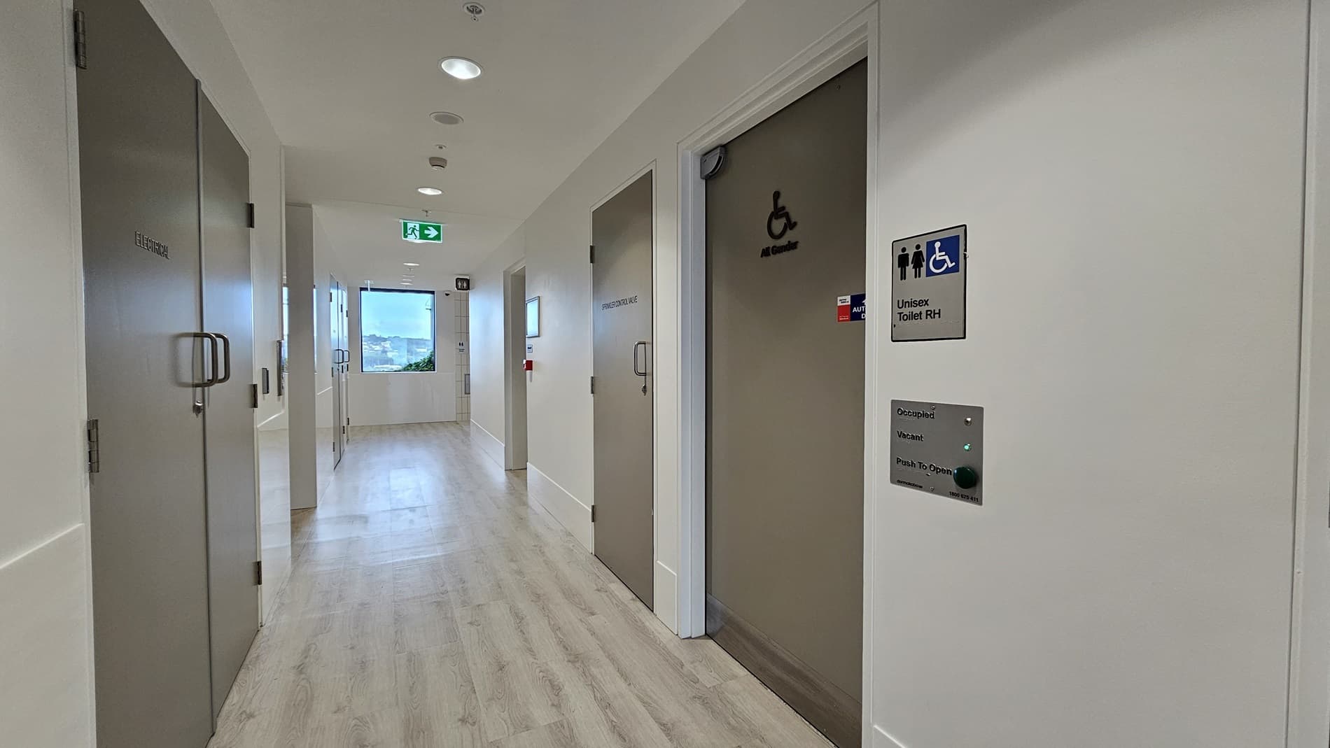 A hallway with a sign on the left that says 'Toilet' with an arrow pointing right and symbols for male, female, and disabled access. There is a door on the left and a doorway leading to another hallway. On the right is a large grey door with a brass strip. A green emergency light is above the doorway to the right. The floor is wood laminate.