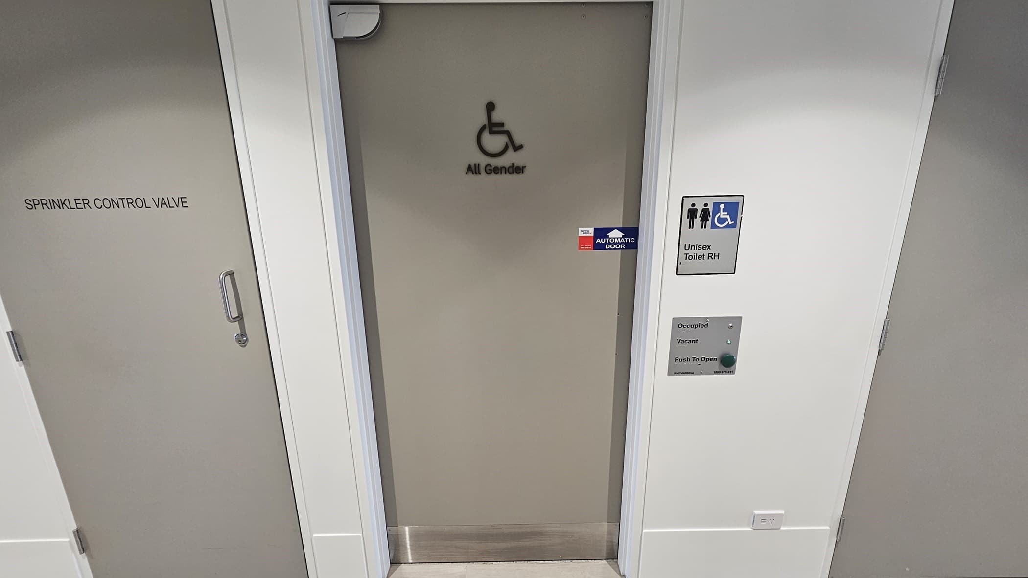 A narrow hallway with white walls and light grey doors. On the left is a door labelled 'SPRINKLER CONTROL VALVE'. The middle door is labelled 'All Gender' and has a wheelchair access symbol. The door on the right is labelled 'Unisex Toilet RH' and has symbols for male, female, and wheelchair access. It also has an indicator showing that it is vacant.