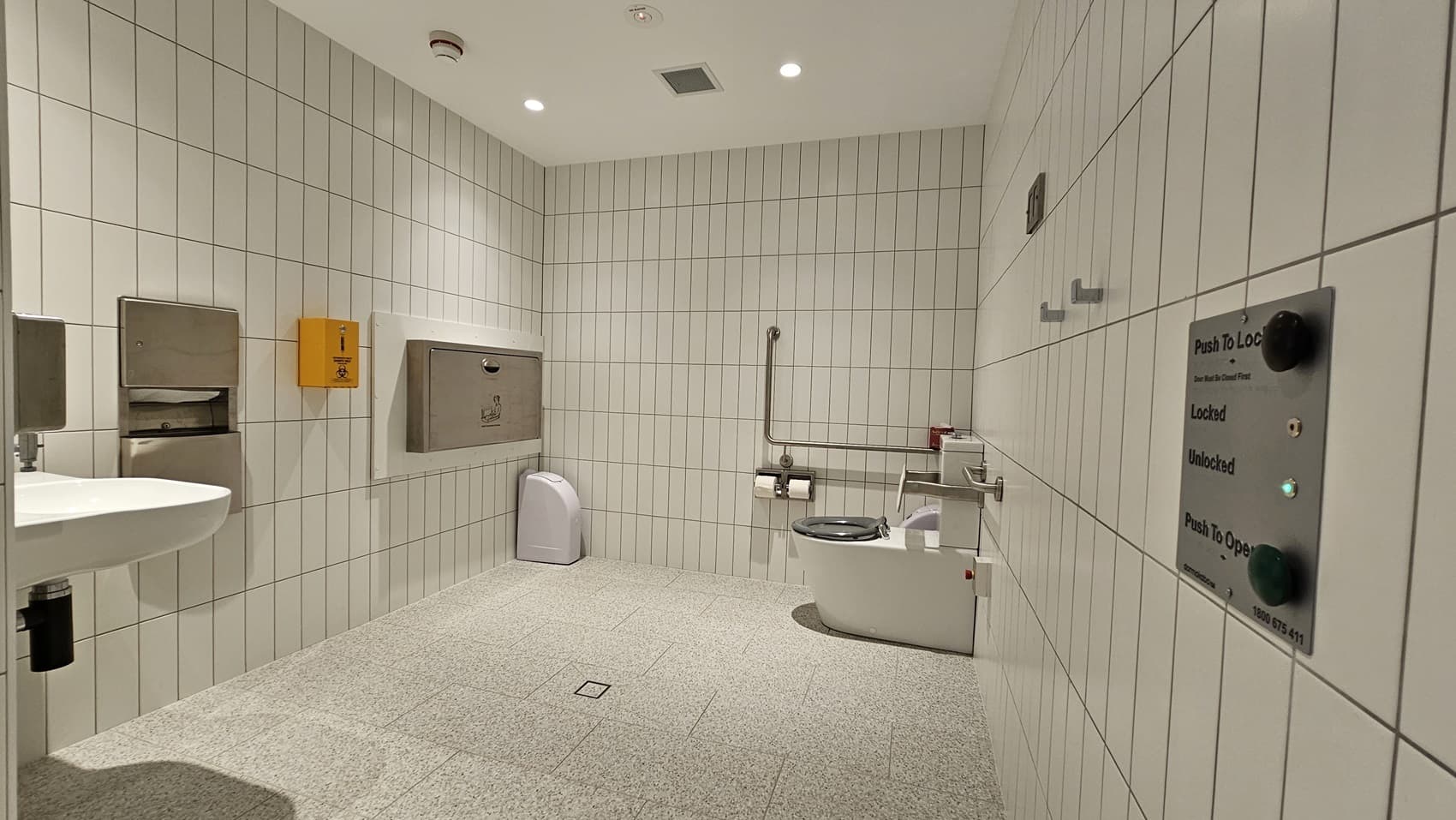 A disabled toilet with white tiled walls and a light grey tiled floor. On the left is a sink with a paper towel dispenser and a hand sanitiser dispenser. On the right is a toilet with grab rails, toilet paper and a control panel to lock and unlock the door. There is also a baby change table on the wall.