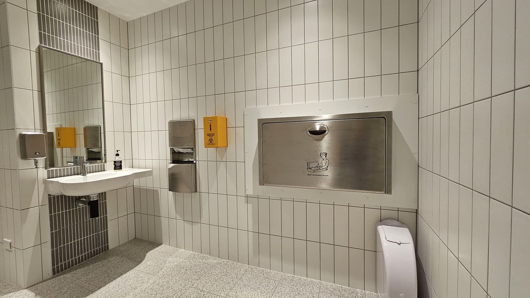 A bathroom with white tiled walls and a light grey tiled floor. On the left is a sink with a mirror behind it and a paper towel dispenser, hand sanitiser dispenser, and liquid soap dispenser. There is a baby change station on the wall on the right.