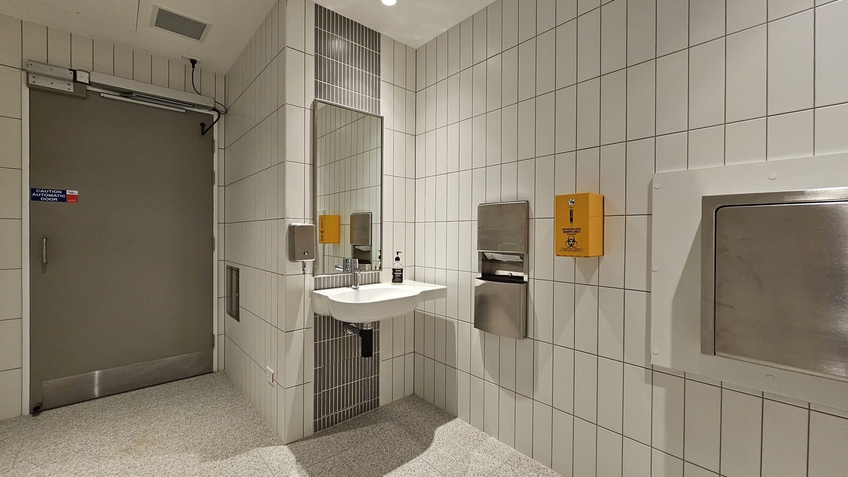 A public washroom with white tiled walls. On the left is a door with a sign that says "CAUTION AUTOMATIC DOOR". On the right is a sink, a mirror, a hand dryer, a hand sanitiser dispenser, and a sharps container. The floor is tiled in a speckled grey pattern.