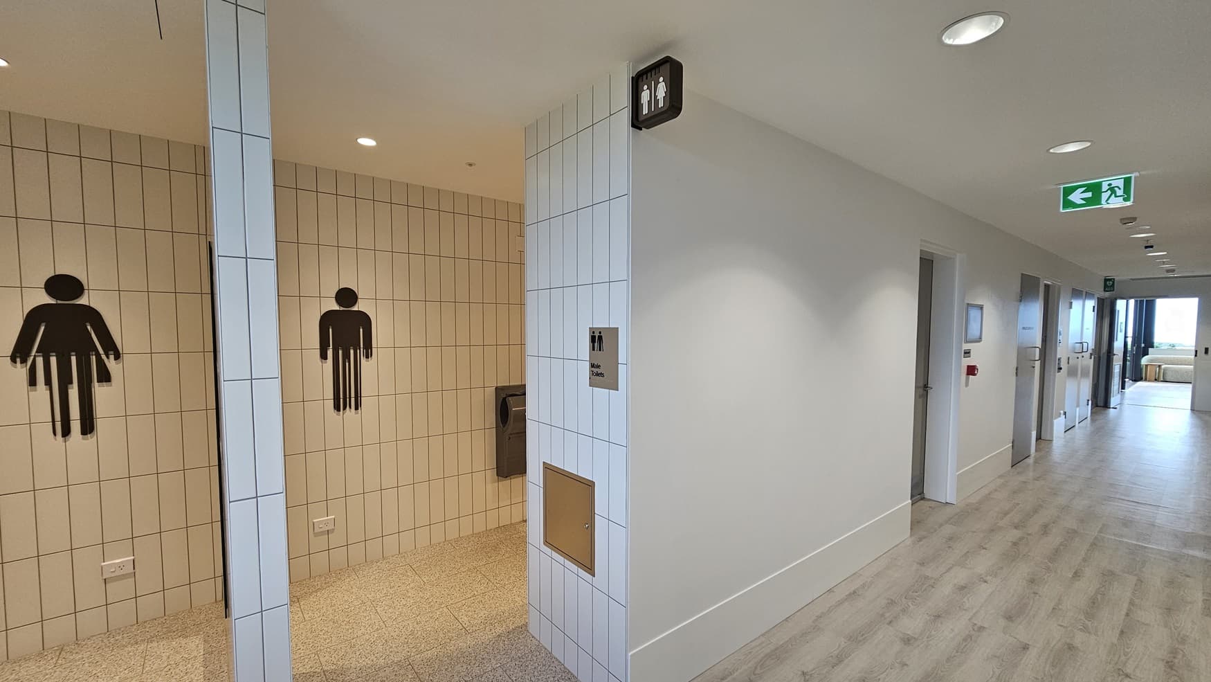 A hallway with white walls and a light wood laminate floor. On the left is the entrance to a toilet with white tiled walls and a sign with pictograms for male and female. The left wall has a hand dryer and an access panel. The hallway leads to a door marked with a green exit sign with a person running to the left. The hallway continues on the right with white doors and a large window at the end.