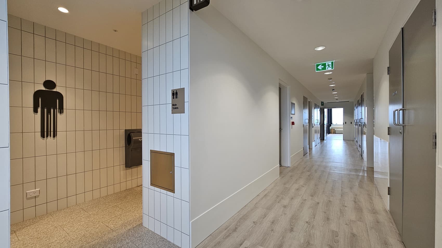 A hallway with white walls and a wood laminate floor. On the left is a tiled wall with a sign that says "Male Toilets" and a silhouette of a man. On the right are a series of doors, the last one is a grey metal door with a silver handle. There is an emergency exit sign with a green arrow pointing left.