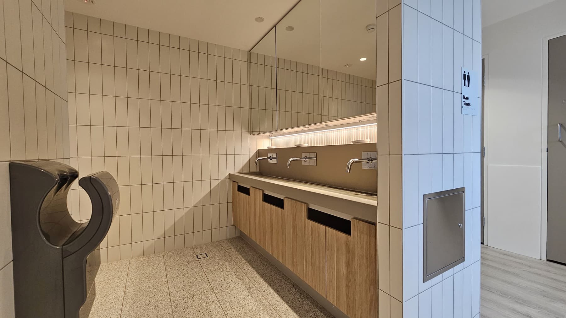 A public washroom with white tiled walls and a speckled grey tiled floor. On the left is a black hand dryer. On the right is a long countertop sink with three chrome taps. Below the sink are wooden cabinets. The wall has a mirror and a sign that reads "Male Toilets".