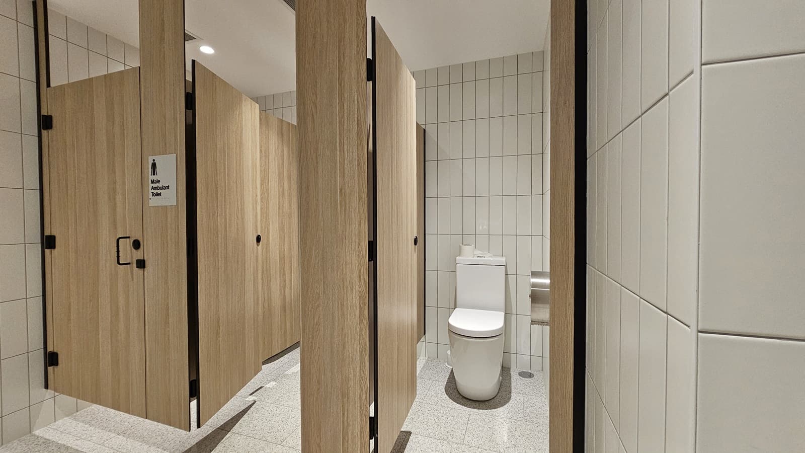 A public toilet cubicle with white tiled walls and a white tiled floor. On the left is a wooden door with a sign that says "Male Ambulant Toilet". There is a toilet on the right with a roll of toilet paper on the back of the toilet.