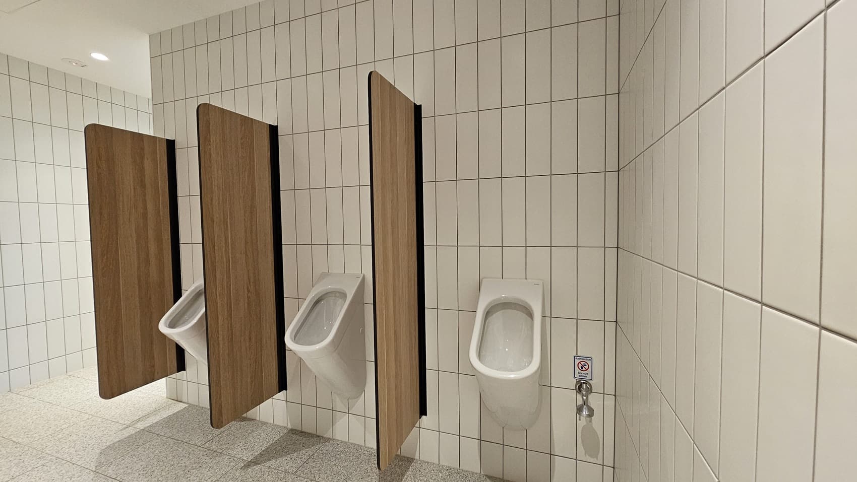 A public washroom with white tiled walls and a speckled grey tiled floor. Four urinals with wooden partitions are mounted on the wall. Each urinal has a chrome flush button. There is a small "no phones" sign on the wall with a phone icon and a red slash through it.