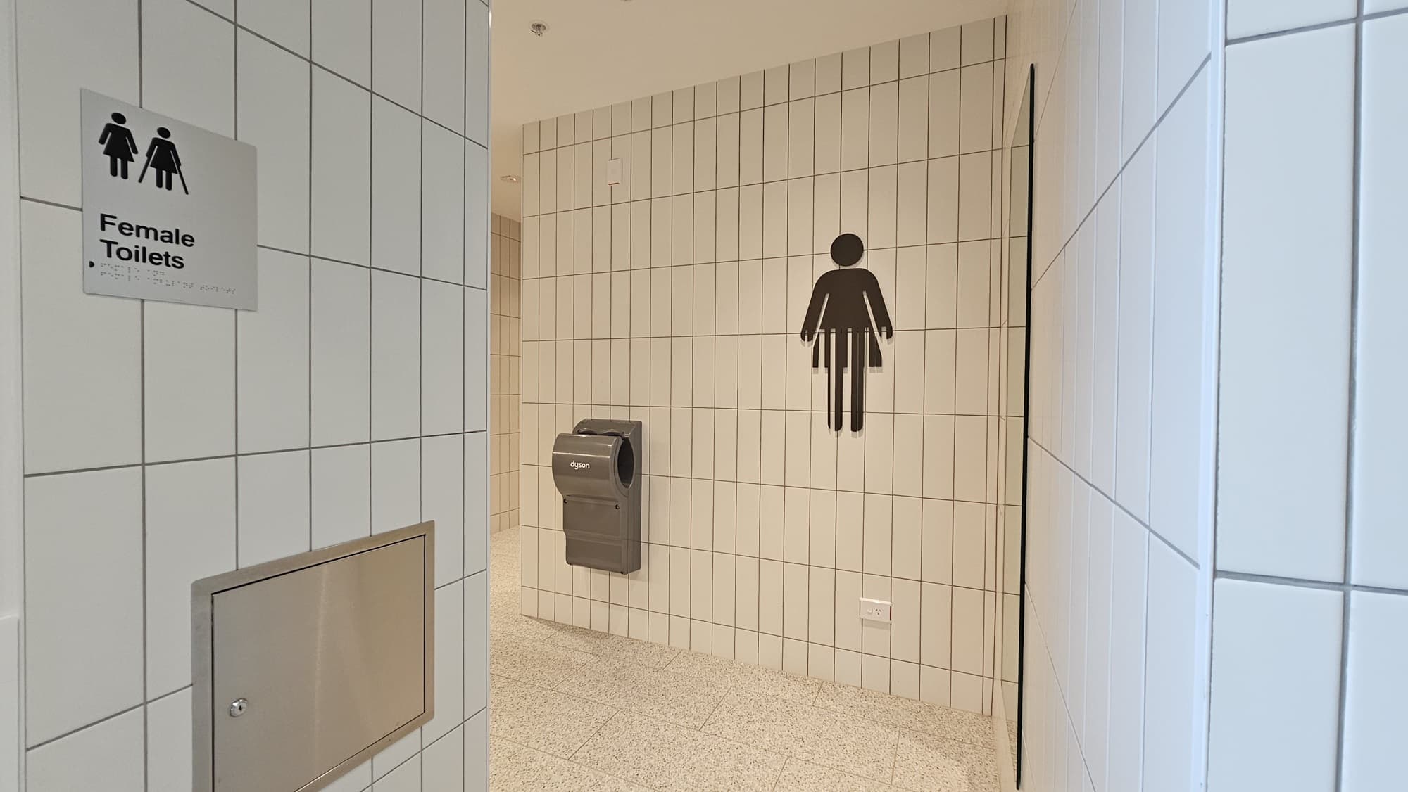 A hallway with white tiled walls and a speckled grey tiled floor. On the left is a door labelled "Female Toilets". It has a black and white sign with two female figures, one with a walking stick. To the right is a grey hand dryer with a sign above it that says "Dyson". A large female figure symbol is on the wall.