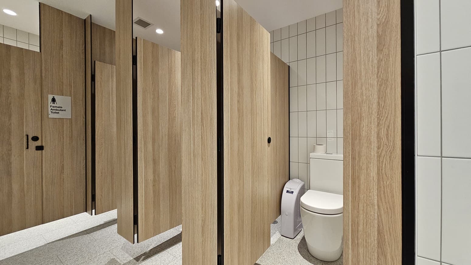A public toilet cubicle with white tiled walls and a speckled grey tiled floor. On the left is a door labelled "Female Ambulant Toilet" with a symbol of a woman in a wheelchair. On the right is a white toilet with a toilet roll holder and a white bin beside it.