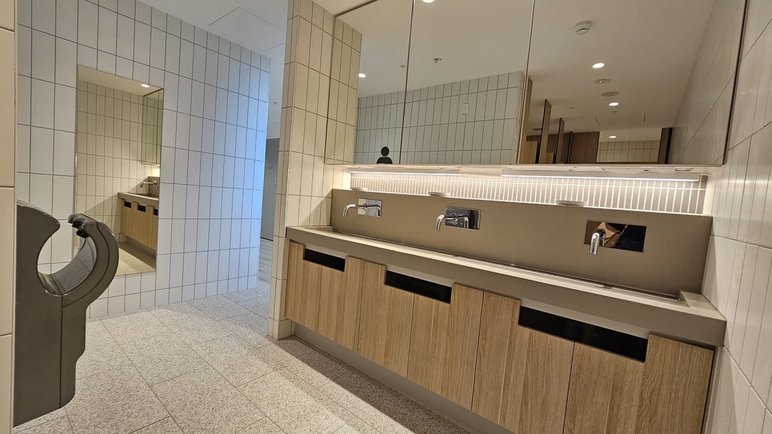 A public washroom with white tiled walls and a speckled grey tiled floor. On the left is a grey hand dryer. On the right is a long countertop sink with two chrome taps, above it is a long mirror. The sink is mounted on a wooden cabinet with three doors and two open shelves.