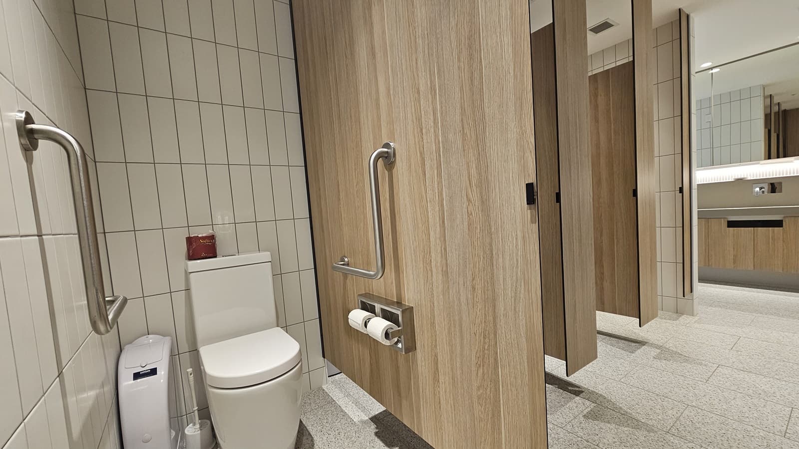 A public toilet cubicle with white tiled walls and a speckled grey tiled floor. On the left is a white toilet with a toilet roll holder, a small bin and a handrail. To the right is a wooden partition with a handrail. There are other cubicles in the background.