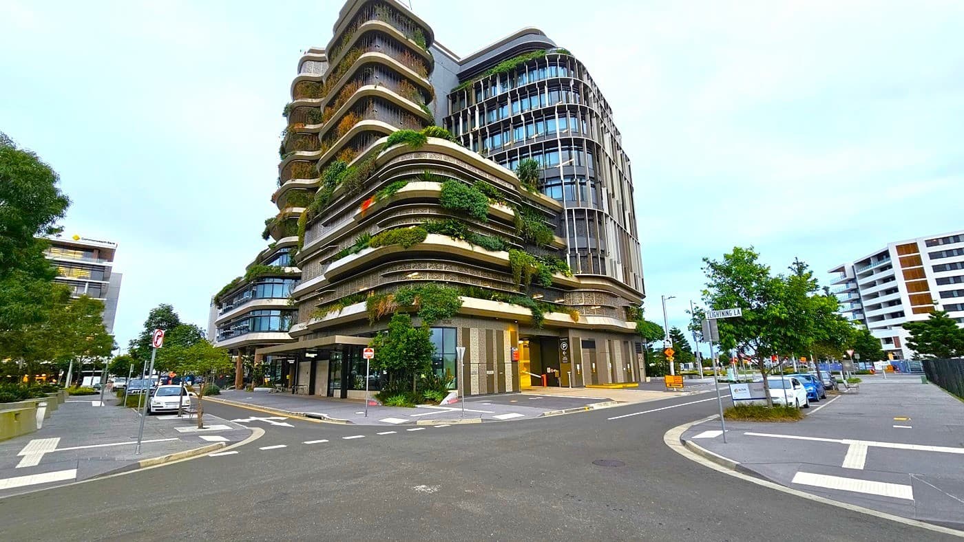 A modern multi-storey building with curved balconies covered in greenery is on the left. On the right, there are smaller buildings and trees lining the street. In the middle, a road intersection with pedestrian crossings and traffic signs. The foreground features a paved road with marked lanes and a few parked cars. The background includes more buildings and trees.