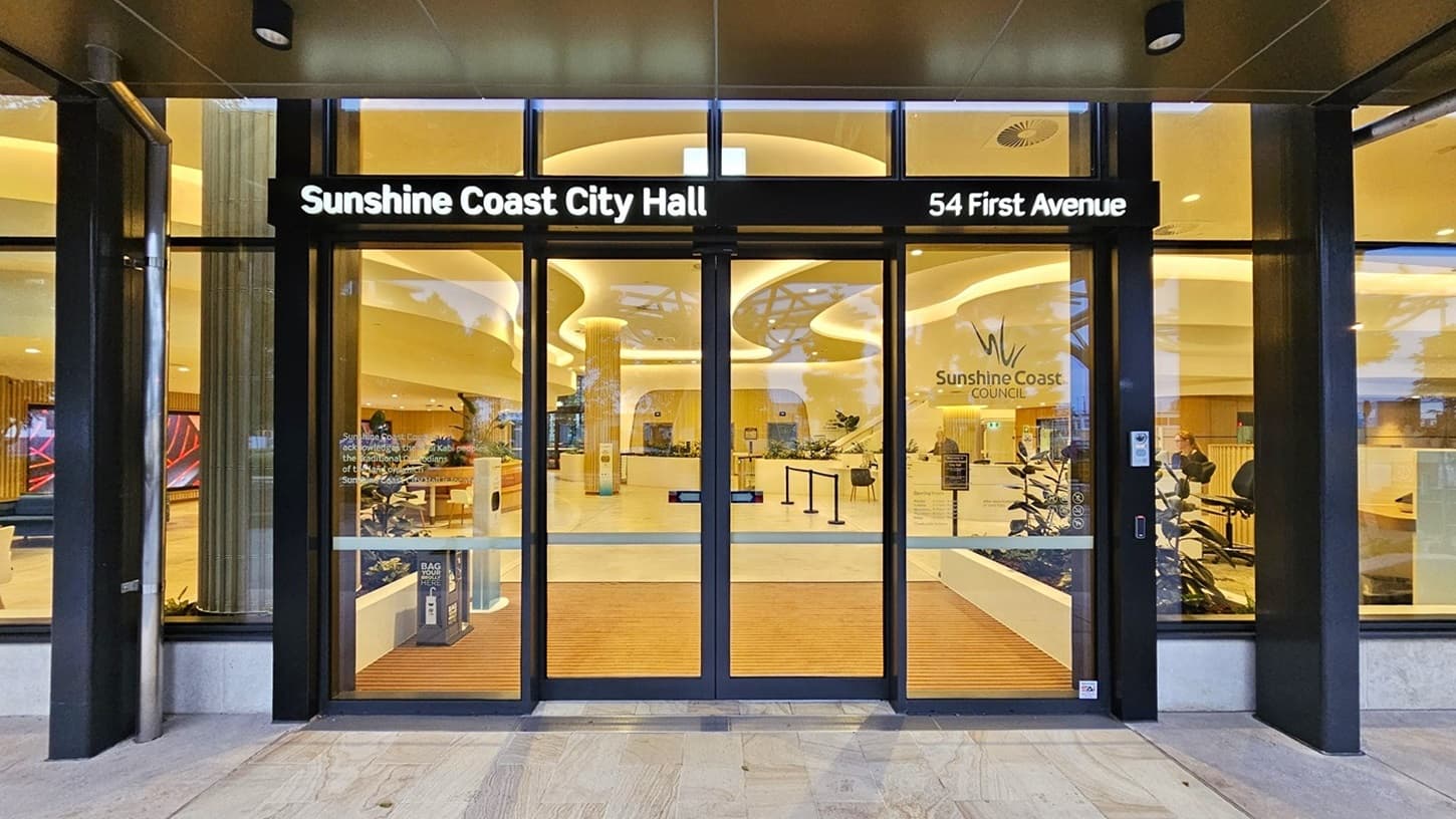 On the left, there is a large glass door with a black frame and a sign reading "Sunshine Coast City Hall." On the right, another glass door with a sign indicating "54 First Avenue." In the middle, the entrance leads to a spacious interior with wooden flooring and modern furnishings. The background features a reception area with a sign for Sunshine Coast Council. The floor outside is light-coloured tiles, and the ceiling inside is curved with recessed lighting.