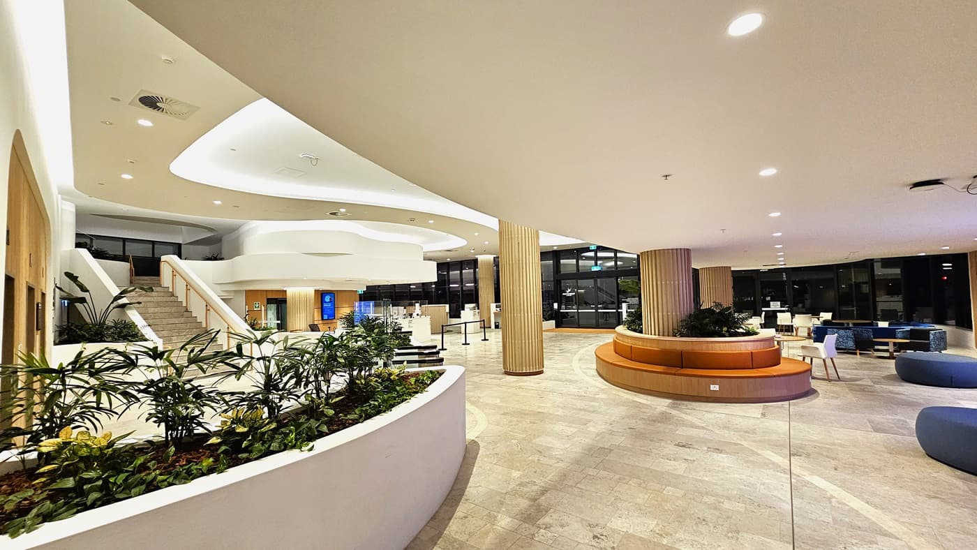 On the left, there is a staircase with wooden handrails leading to an upper level, flanked by a white curved wall with potted plants. On the right, there are circular seating areas with orange cushions and a few blue chairs. In the middle, a large open space with light-coloured tiled flooring and several wooden columns. The background features large windows and more seating areas. The ceiling is white with recessed lighting and curved architectural elements.