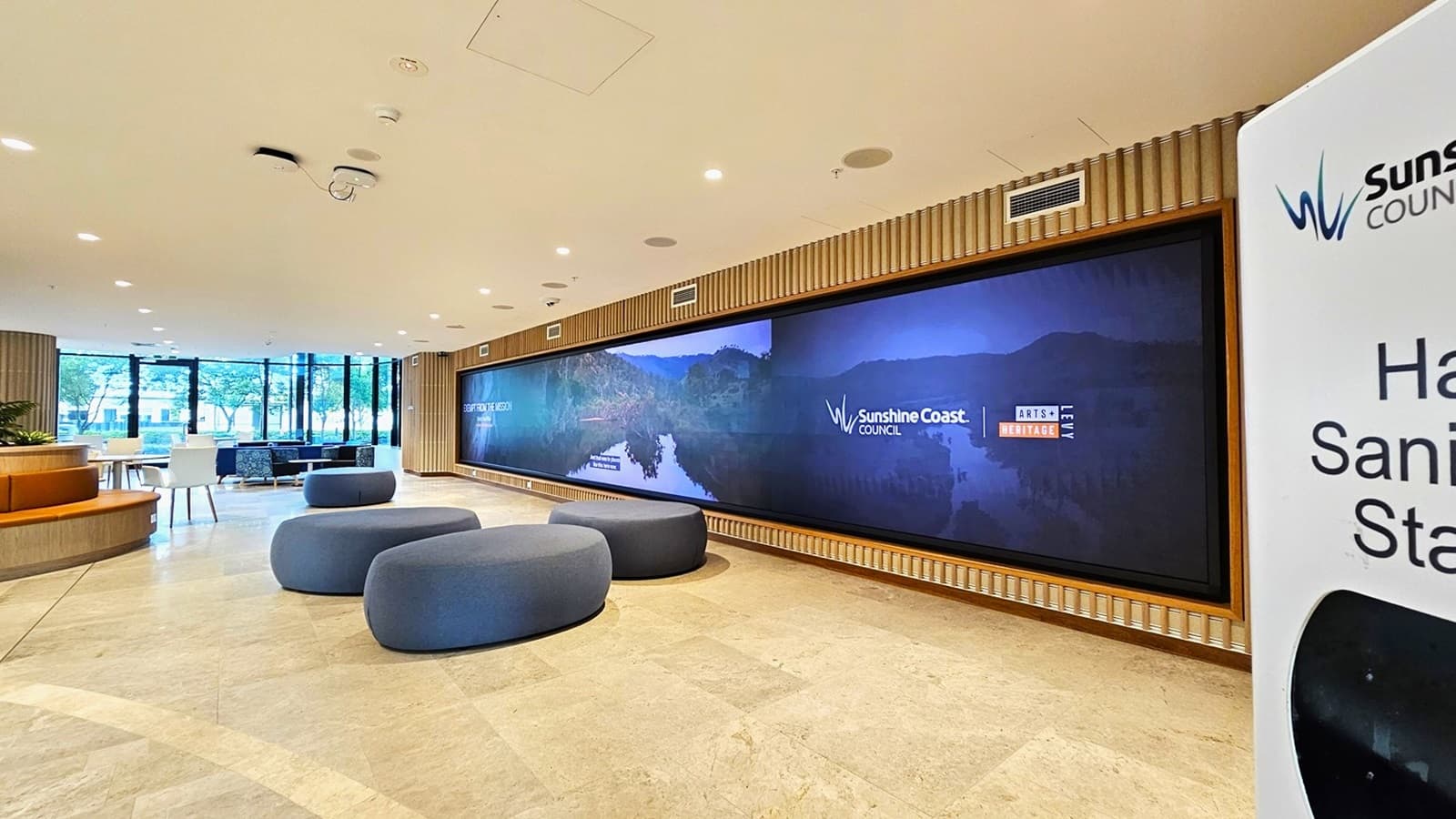 On the left, there are round blue seating poufs and a curved wooden bench with a few potted plants. On the right, a large banner with the Sunshine Coast Council logo and a hand sanitiser station. In the middle, a long digital display screen showing scenic images and the Sunshine Coast Council logo. The background features large windows with a view of trees and more seating. The floor is light-coloured tiles, and the ceiling is white with recessed lighting and ventilation panels.