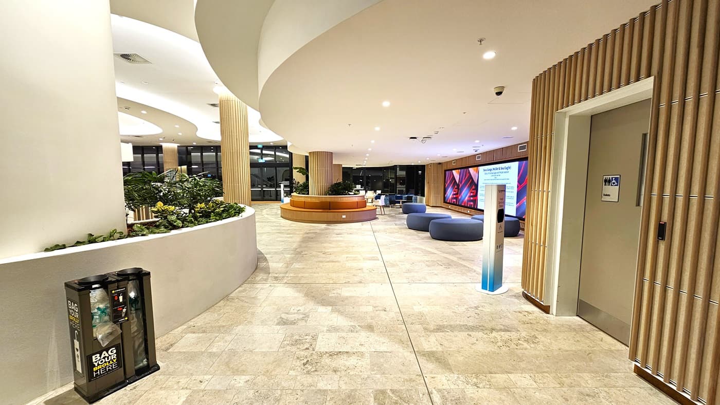 On the left, there is a curved white wall with a planter containing various green plants and a recycling bin. On the right, there is a lift with a sign indicating "Lifts" and a wooden panelled wall. In the middle, there are blue circular seating poufs and a curved wooden bench. The background features a large digital display screen and more seating. The floor is light-coloured tiles, and the ceiling is white with recessed lighting and curved architectural elements.