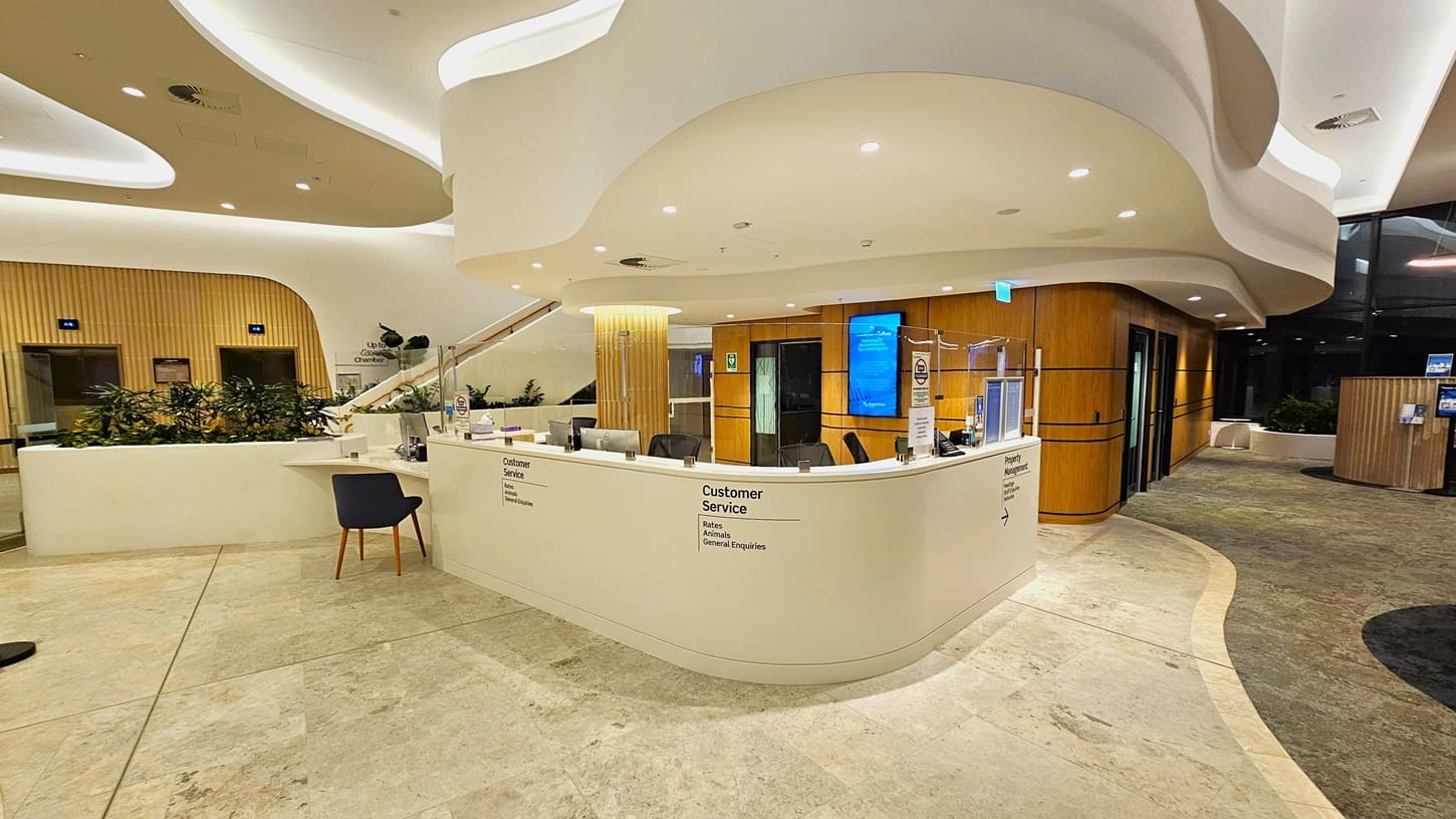 On the left, there is a curved white counter with a "Customer Service" sign, a blue chair, and potted plants behind it. On the right, there is a wooden panelled wall with a digital screen and a lift. In the middle, the counter extends with more signage and a computer monitor. The background features more wooden panelling, a digital display, and a glass wall. The floor is light-coloured tiles, and the ceiling is white with recessed lighting and curved architectural elements.