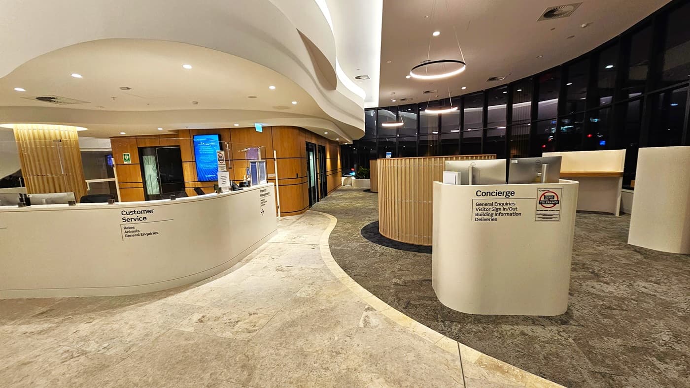 On the left, there is a curved white counter with a "Customer Service" sign, a blue chair, and potted plants behind it. On the right, there is a smaller curved counter labelled "Concierge" with signs in multiple languages. In the middle, the floor transitions from light-coloured tiles to darker tiles near the counters. The background features large floor-to-ceiling windows with black frames and a view of the outside. The ceiling is white with recessed lighting and curved architectural elements.