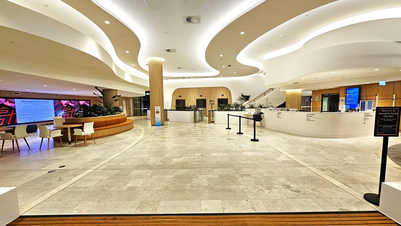 On the left, there is a curved wooden bench with white and blue chairs around small tables, and a digital display screen with vibrant colours behind it. On the right, there is a long, curved white counter with "Customer Service" and "Concierge" signs, along with potted plants and a few computer monitors. The background features more seating, wooden columns, and large windows. The ceiling is white with recessed lighting and curved architectural elements.