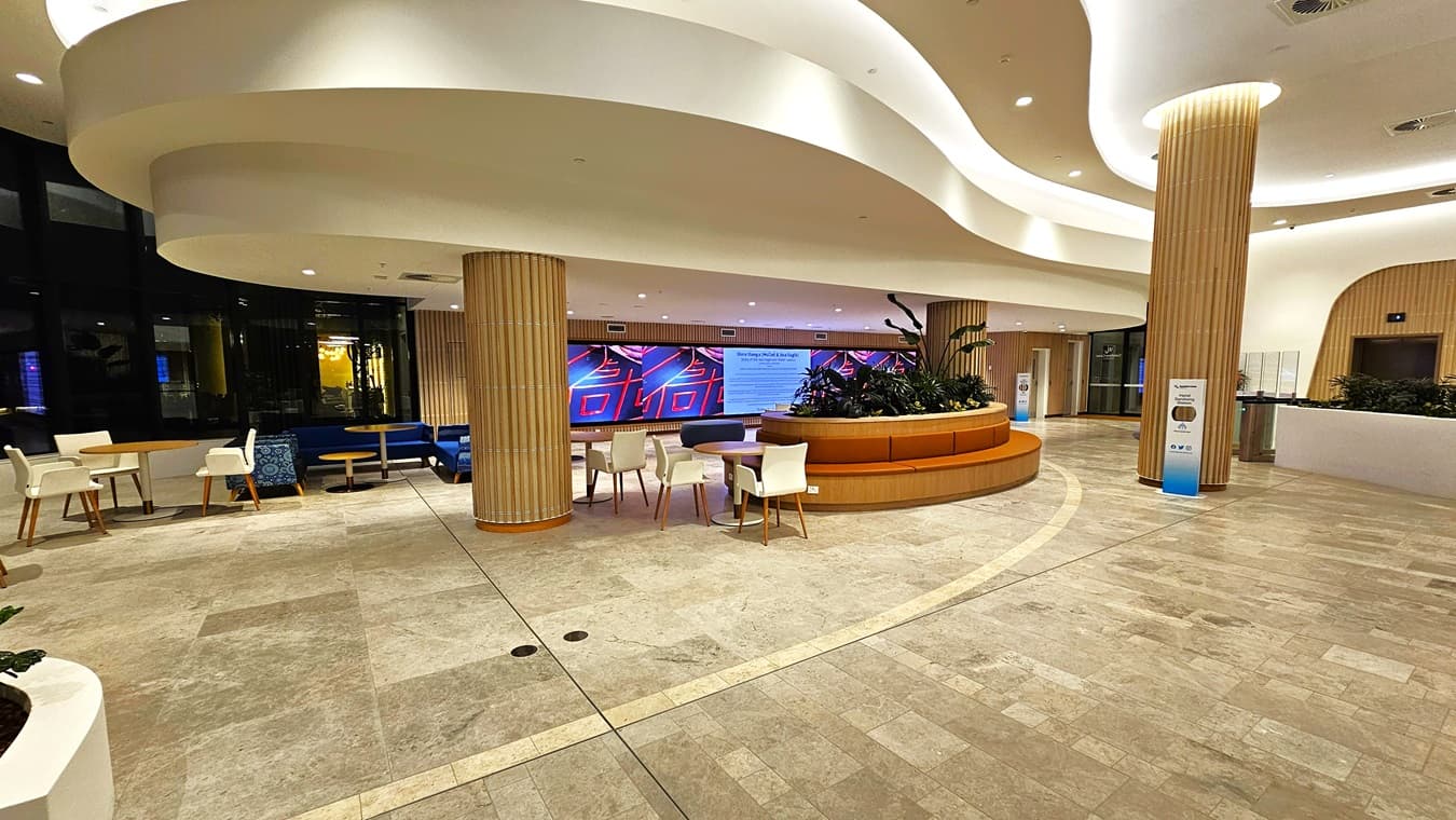 On the left, there are white and blue chairs around small tables, with a curved wooden bench and a digital display screen showing vibrant colours behind it. On the right, there is a long, curved white counter with potted plants and a sign. In the middle, there is a spacious area with light-coloured tiled flooring and a circular wooden seating area with plants. The ceiling is white with recessed lighting and curved architectural elements.