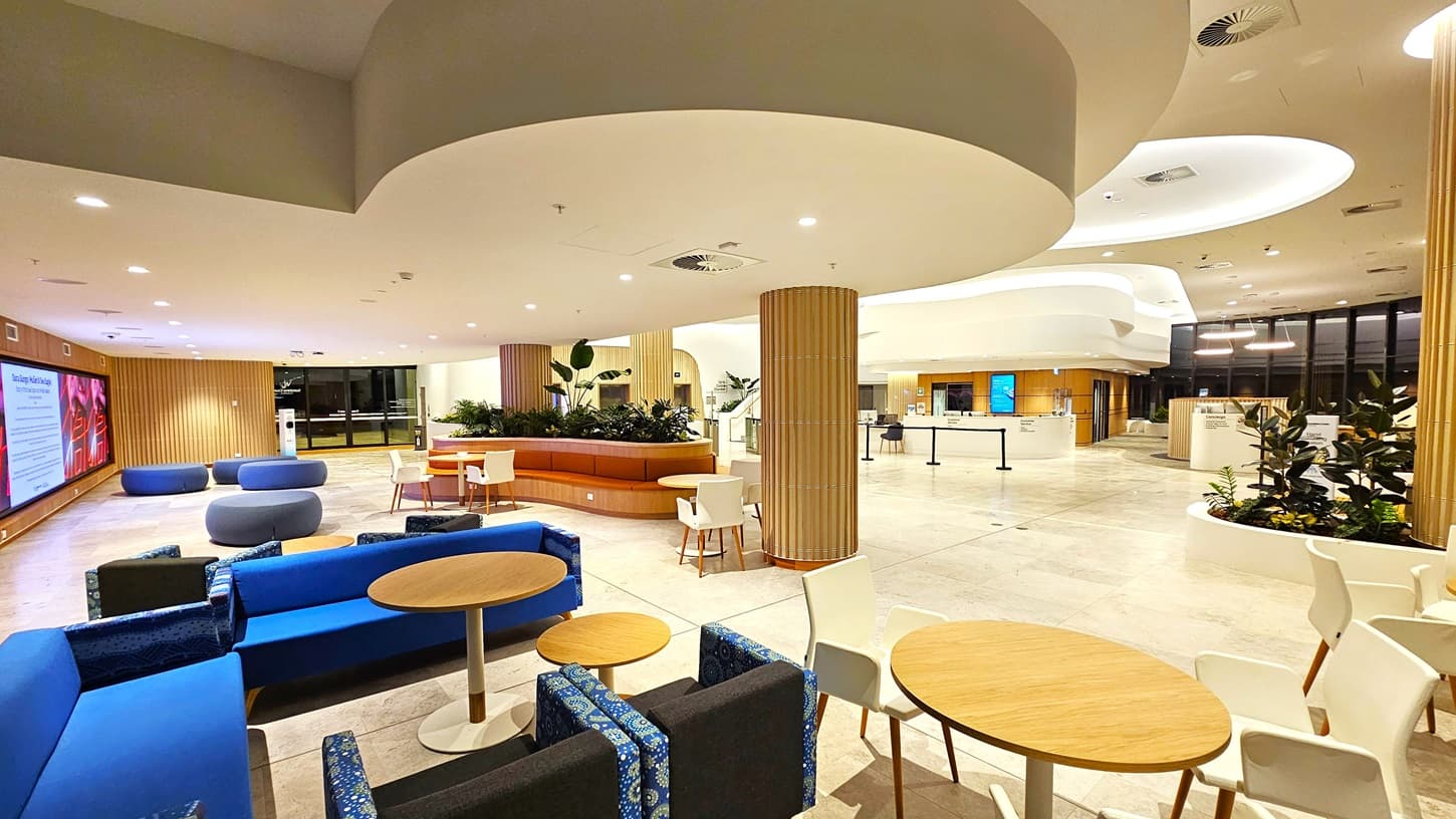 On the left, there are blue and grey sofas with wooden tables, and a large digital display screen on the wall. On the right, there is a white counter with potted plants and a sign, along with more seating and tables. In the middle, there is a circular wooden seating area with plants. The background features large windows, additional seating, and a white counter. The floor is light-coloured tiles, and the ceiling is white with recessed lighting and curved architectural elements.