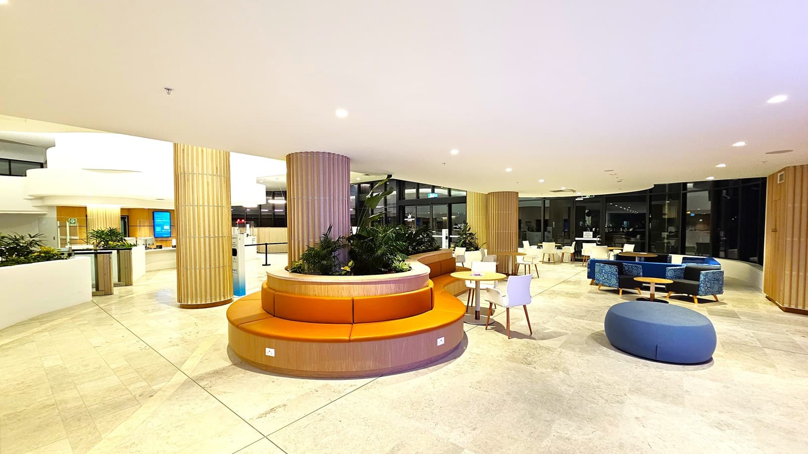 On the left, there is a white counter with potted plants and a digital display screen in the background. On the right, there are blue and white chairs around small tables, and a blue circular seating pouf. In the middle, there is a circular wooden seating area with orange cushions and green plants in the centre. The background features large windows, additional seating, and wooden columns. The floor is light-coloured tiles, and the ceiling is white with recessed lighting.