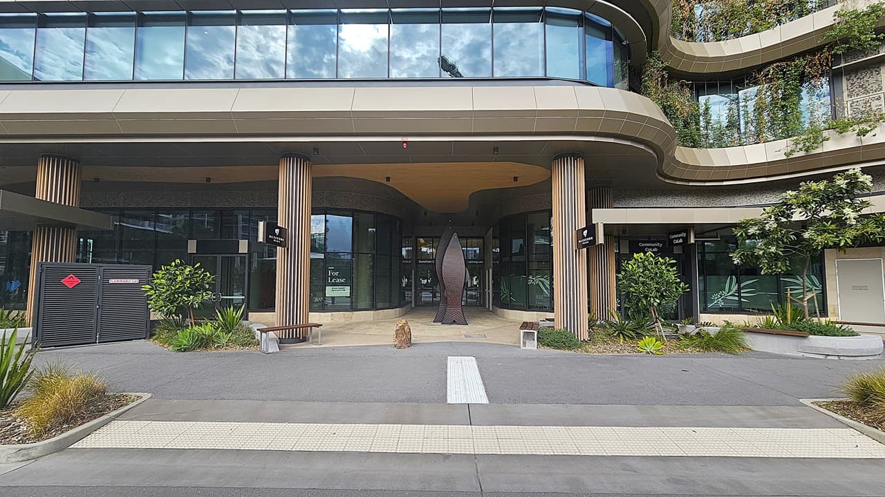 A modern building entrance with a grey paved walkway and a beige tiled strip in the foreground. On the left, there is a black utility box and a wooden bench. In the middle, there is a large glass entrance with wooden columns and a tall, abstract sculpture. The ceiling above the entrance is beige with recessed lighting. On the right, there are more wooden columns, green plants, and a door. The background features large windows and greenery on the upper levels.