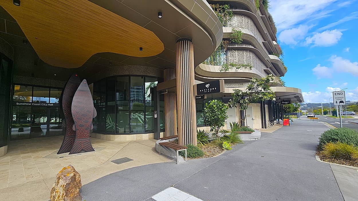A modern building entrance with a grey paved walkway and beige tiles in the foreground. On the left, there is a large glass door with "Sunshine Coast City Hall" written on it, and a tall, abstract sculpture next to a wooden bench. The ceiling above the entrance is beige with recessed lighting. On the right, there are wooden columns, green plants, and a sign for "Giddy Geisha." The background features a curved building facade with greenery on the balconies and a road with a traffic sign.