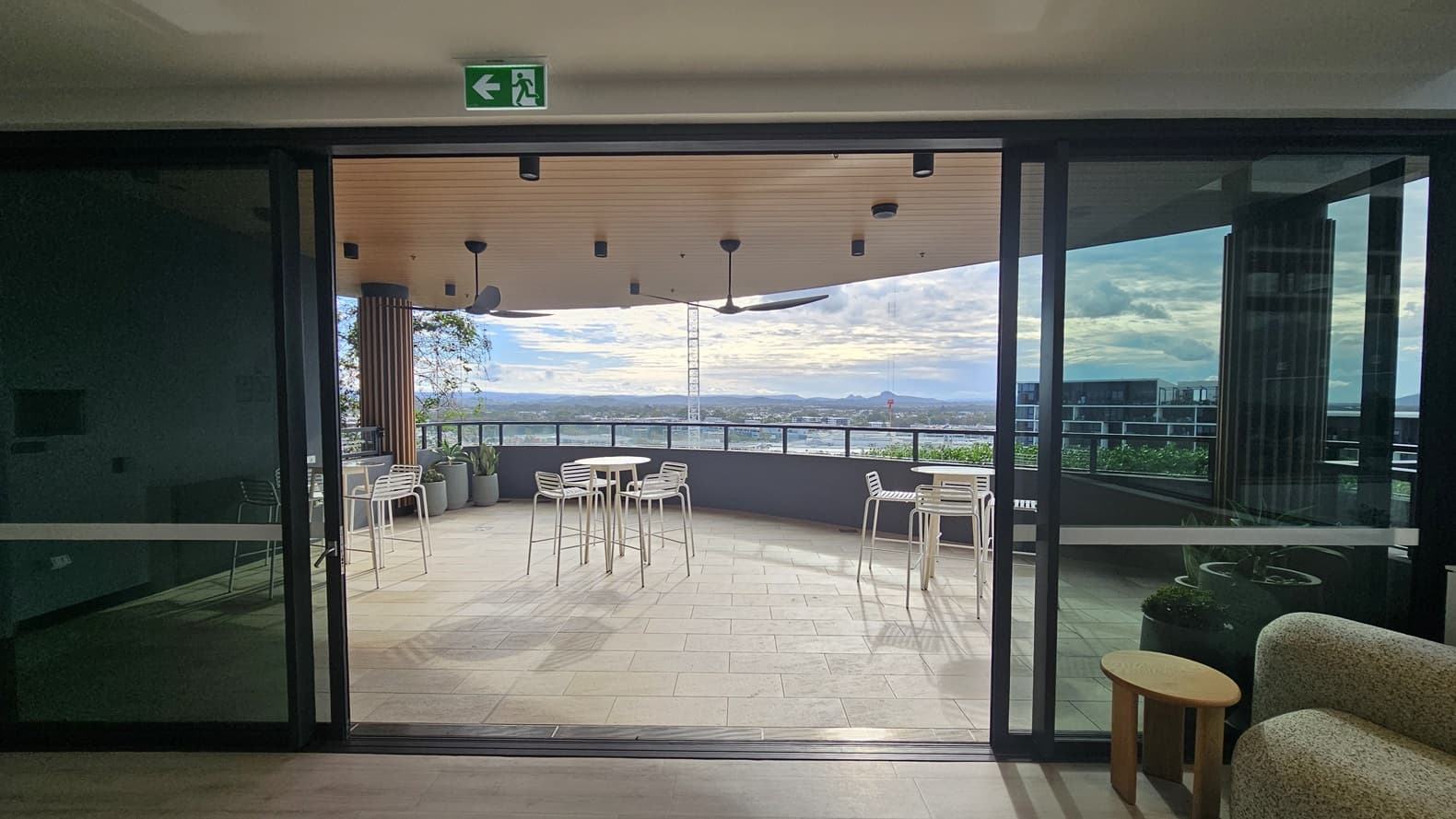 A set of sliding glass doors leading to an outdoor balcony. The balcony has a light coloured tiled floor, a grey railing, a wooden ceiling and a few tables and chairs. In the background, there is a view of a city with buildings and a distant mountain.