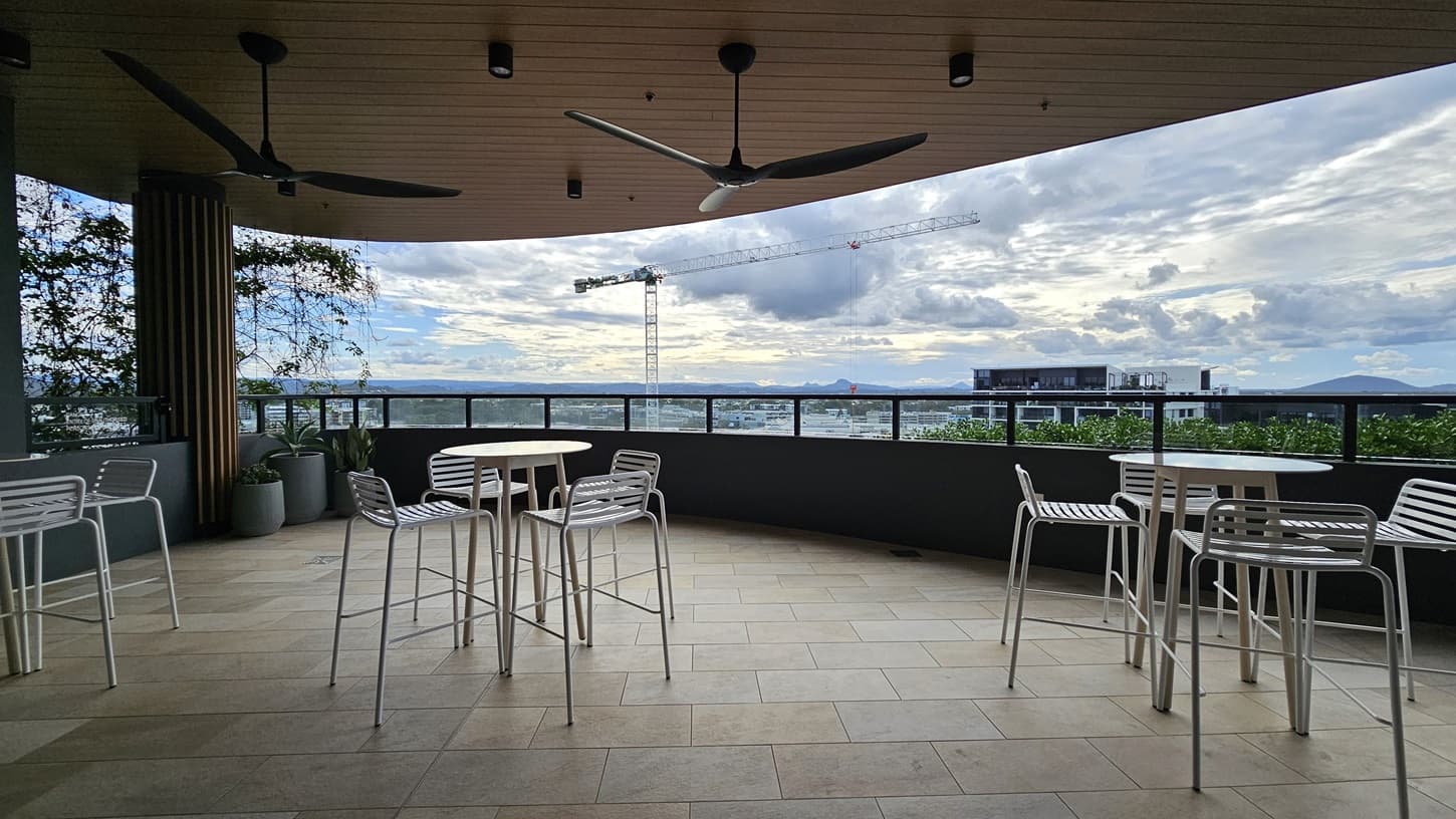 On the left, there are two white tables with white chairs and potted plants near a wooden ceiling with ceiling fans. On the right, there are two more white tables with white chairs. In the middle, the balcony has a tiled floor and a railing overlooking a cityscape with buildings and a construction crane in the background.