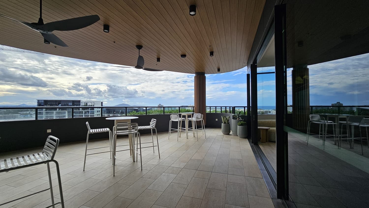 On the left, there are two white tables with white chairs and a ceiling fan attached to a wooden ceiling. On the right, there is a glass wall reflecting the balcony and a black railing. In the middle, the balcony has a tiled floor and potted plants near the railing. In the background, there are buildings and a cloudy sky.
