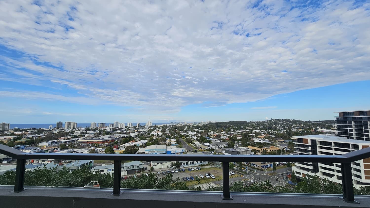On the left, there are several low-rise buildings and a few taller structures in the distance. On the right, there is a modern multi-storey building with white and black accents. In the middle, the view extends over a sprawling urban area with roads, houses, and commercial buildings. The foreground features a black railing.