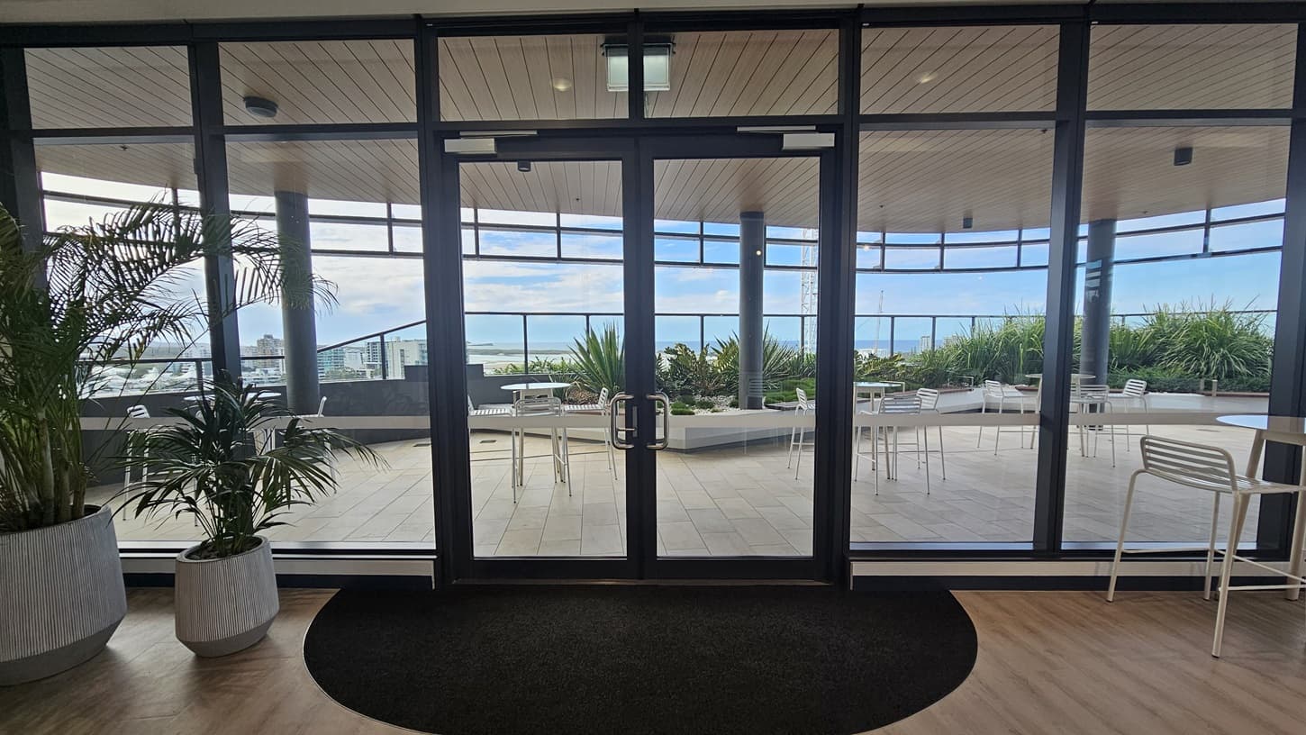 On the left, there are potted plants near a glass door. On the right, there are white tables and chairs next to the glass wall. In the middle, the glass doors open to a balcony with more white tables and chairs. The background features greenery and a distant view of buildings. The floor inside is wooden, transitioning to tiled on the balcony, and the ceiling is wooden with recessed lighting.
