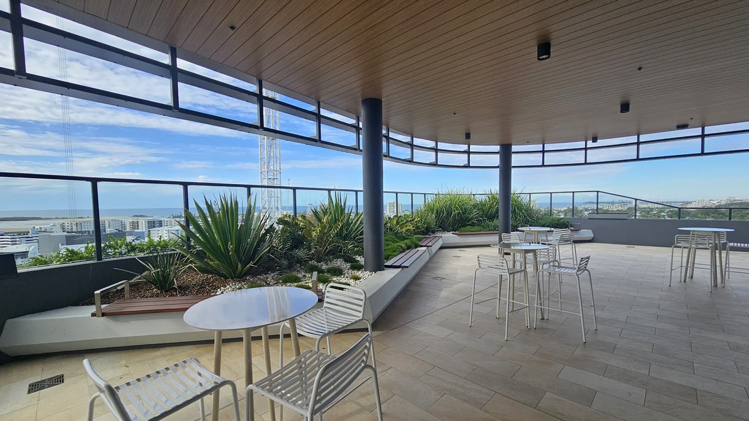 On the left, there are white tables and chairs near a glass railing with a view of buildings and a crane in the distance. On the right, there are more white tables and chairs. In the middle, there are large potted plants and a bench along the glass railing. The background features more buildings and greenery. The floor is tiled, and the ceiling is wooden with recessed lighting.