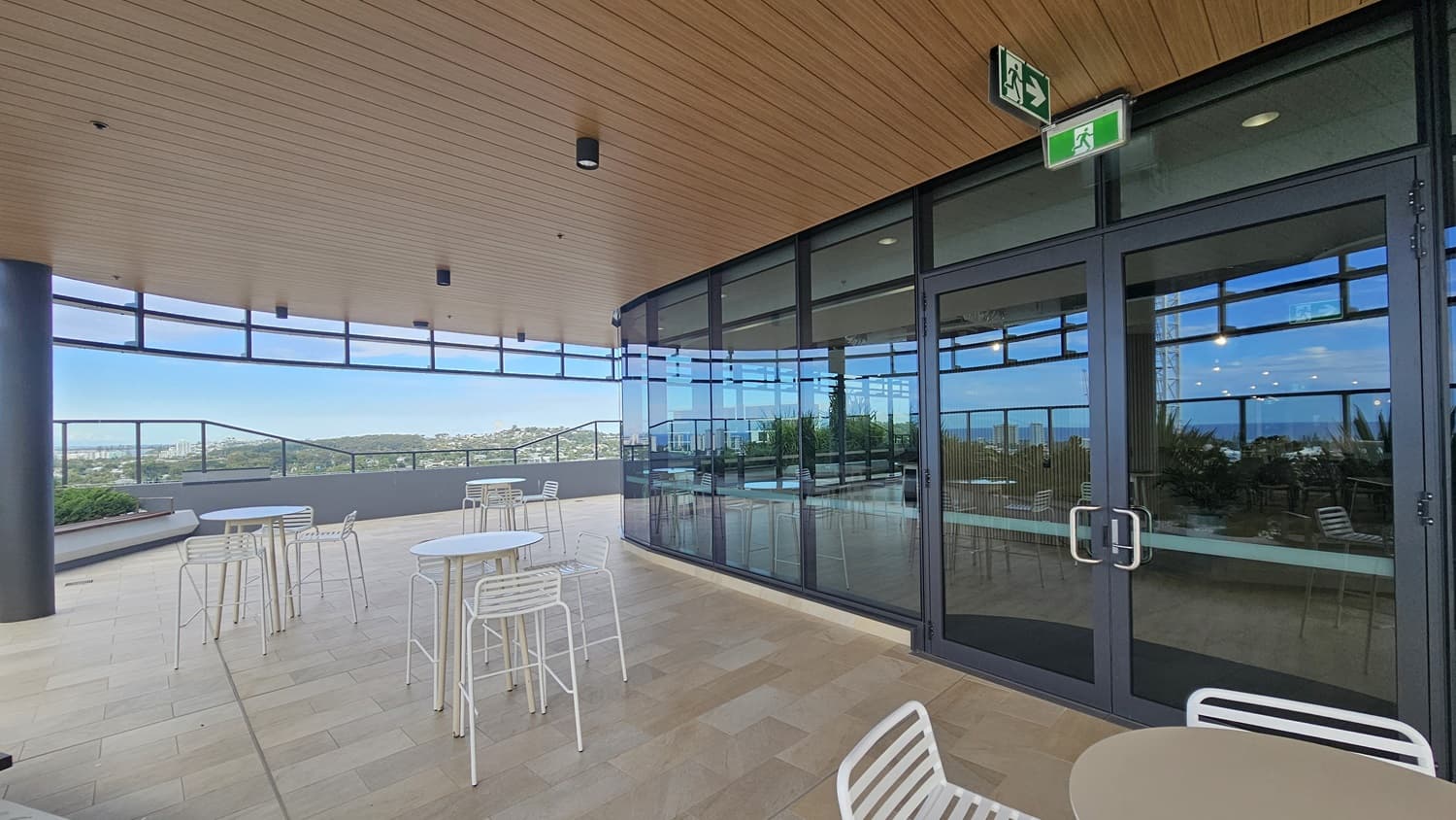 On the left, there are white tables and chairs on a tiled floor near a glass railing overlooking a distant landscape. On the right, there is a glass wall with double doors leading to an indoor area. In the middle, more white tables and chairs are arranged on the balcony. The background features a view of hills and buildings. The ceiling is wooden with recessed lighting and an emergency exit sign.