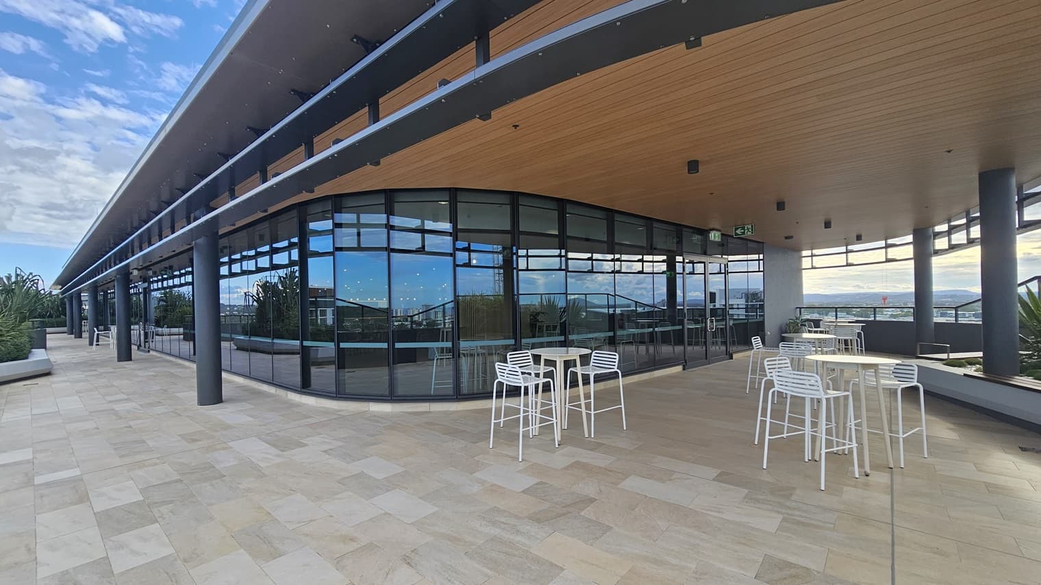 On the left, there are potted plants and a long bench along the edge of the balcony. On the right, there are white tables and chairs on a tiled floor. In the middle, there is a curved glass wall with black frames and reflections of the surroundings. The background includes more seating and a view of distant hills and buildings. The ceiling is wooden with recessed lighting and black support beams.