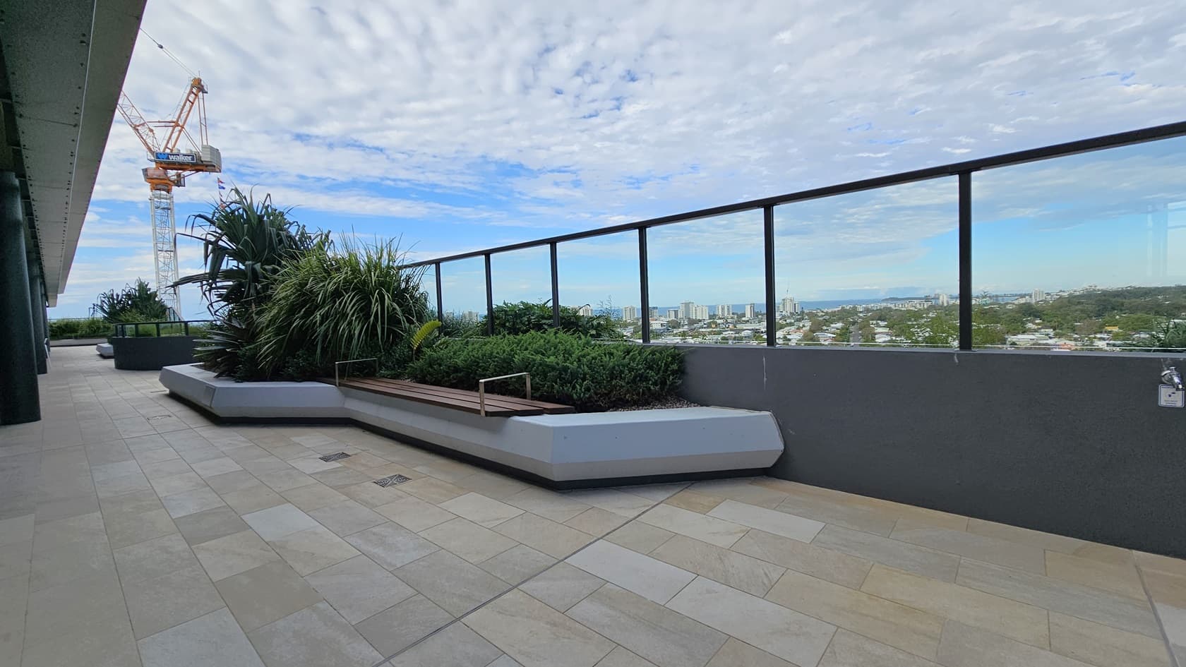 On the left, there is a large planter with tall green plants and a construction crane in the distance. On the right, there is a glass railing with a view of a cityscape and distant buildings. In the middle, there is a long bench integrated into a planter with various green shrubs. The floor is tiled with light-coloured stone tiles.