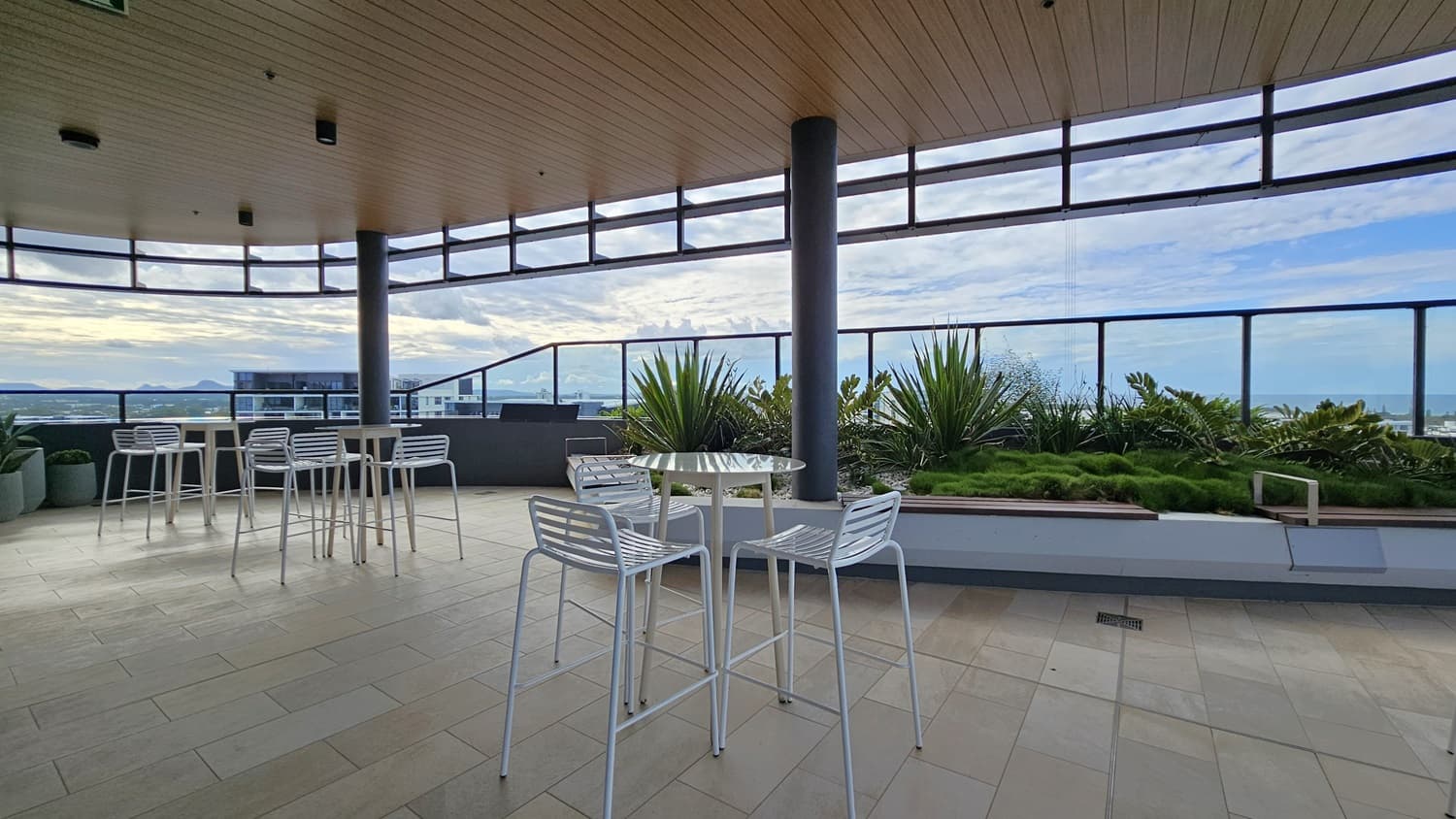 On the left, there are white high tables and chairs on a tiled floor, with a glass railing and a view of distant buildings. On the right, there is a raised planter with green shrubs and a glass railing overlooking the landscape. In the middle, there are more white high tables and chairs. The floor is tiled with light-coloured tiles, and the ceiling is wooden with recessed lighting and black support beams.