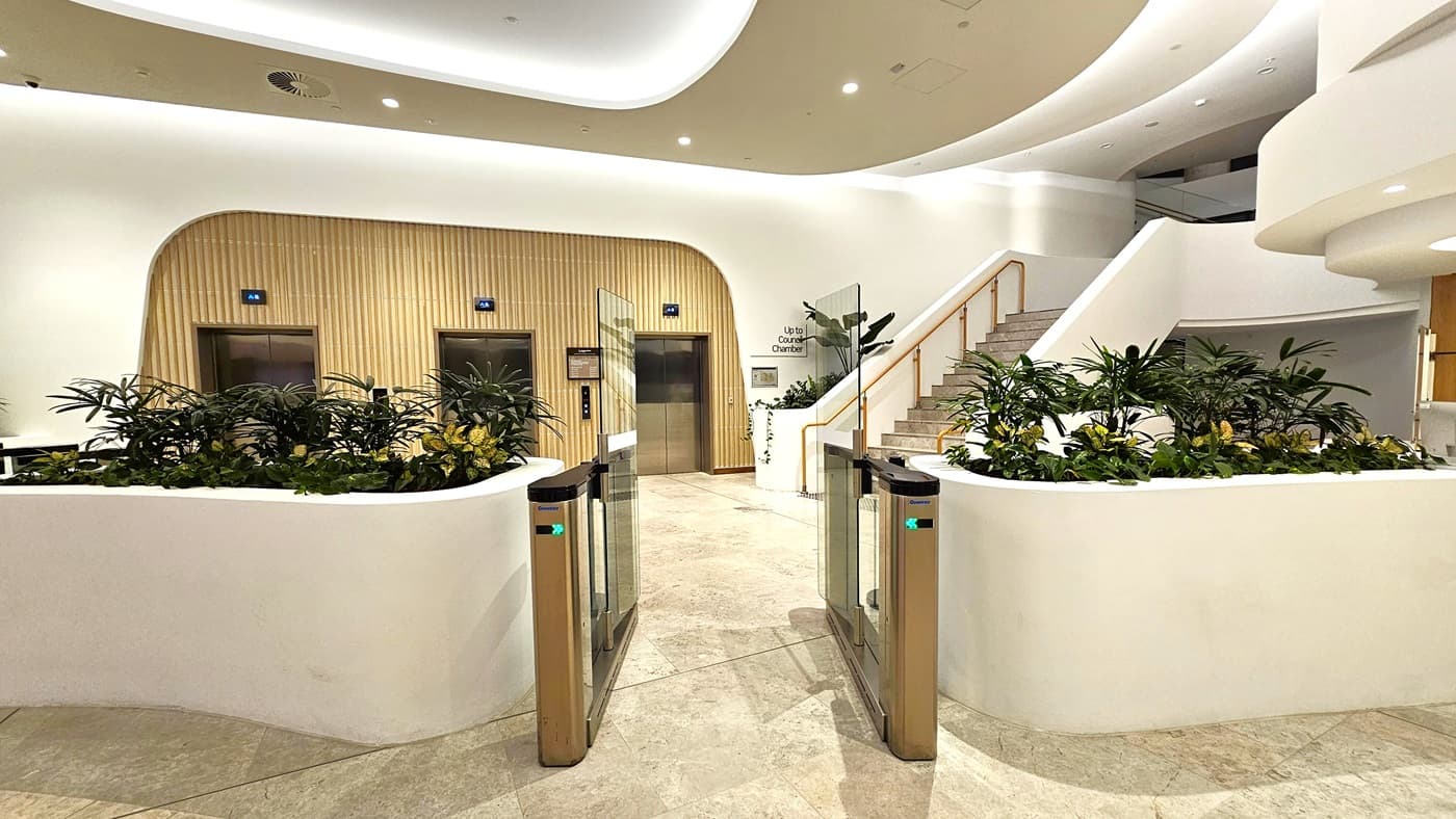 On the left, there are two lifts with wooden panelling and blue indicators above them, surrounded by potted plants. On the right, there is a staircase with a wooden handrail leading to an upper level, also surrounded by potted plants. In the middle, there is a gated entrance with glass barriers and green indicator lights. The background features plants and a white wall. The floor is light-coloured marble tiles, and the ceiling is white with recessed lighting and curved architectural elements.