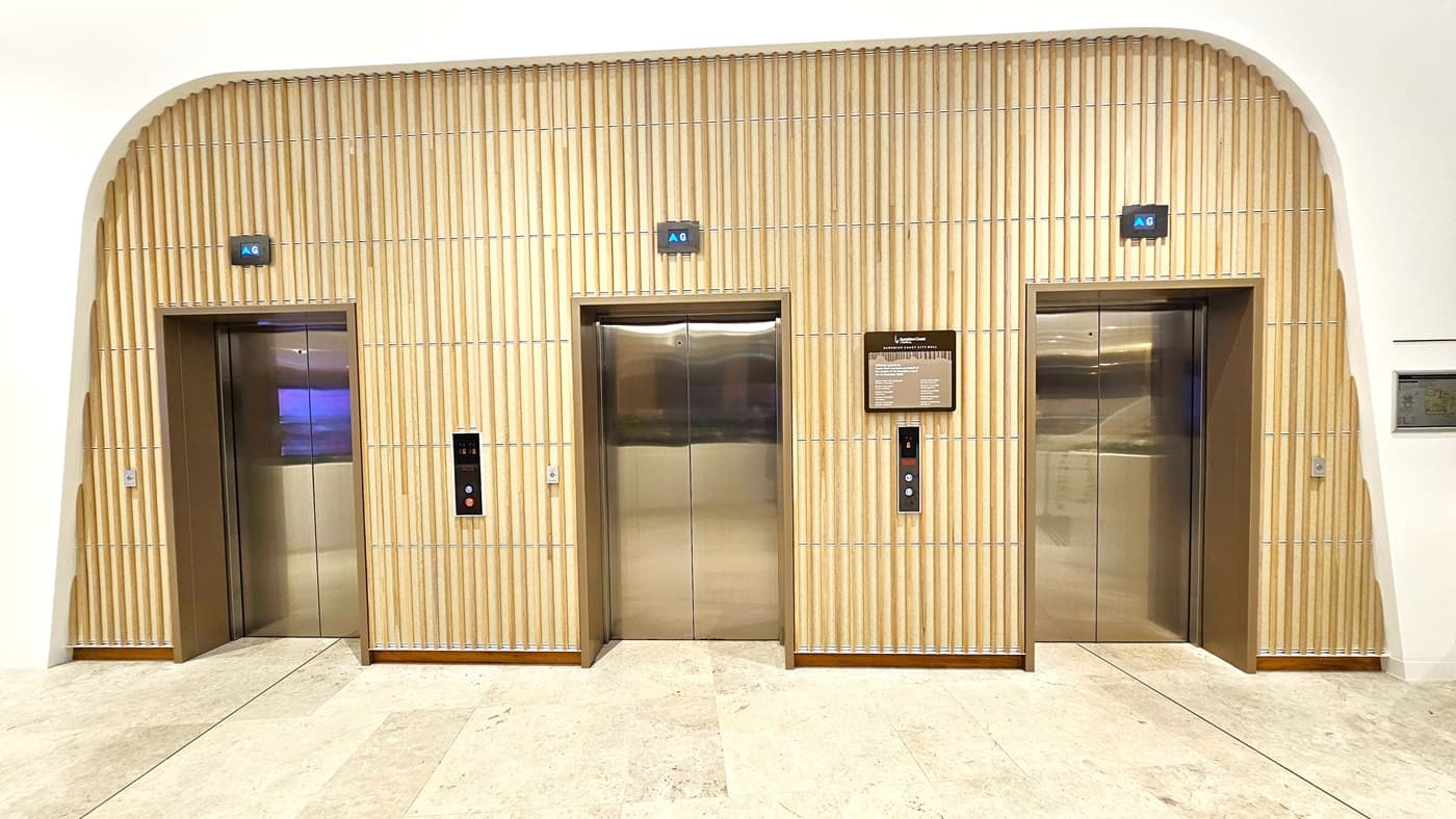 On the left, there is a lift with a blue indicator above it, set within a wooden panelled wall. On the right, there is another lift with a blue indicator above it, also within the same wooden panelled wall. In the middle, there is a third lift with a blue indicator above it, and a sign displaying floor information. The floor is light-coloured marble tiles, and the ceiling is white with a curved edge where it meets the wooden panelling.
