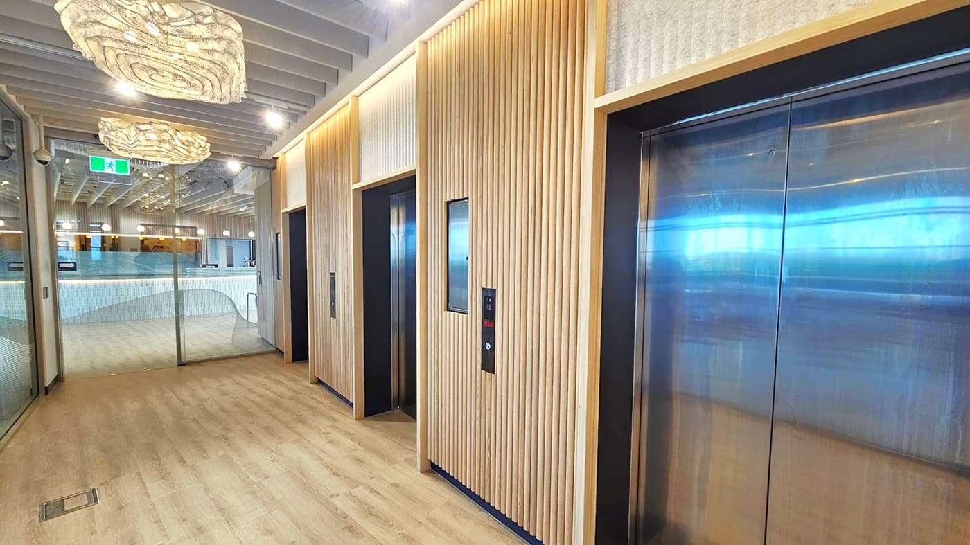 A hallway with three sets of elevator doors on the right. The doors are made of stainless steel and have a black frame. There is a wood panelled wall behind the doors. The ceiling has light wood panels and two large, light-coloured, organic shaped lights hanging from it. The floor is a light brown wood laminate. On the left there is a glass door leading into a room. The floor is light wood laminate.