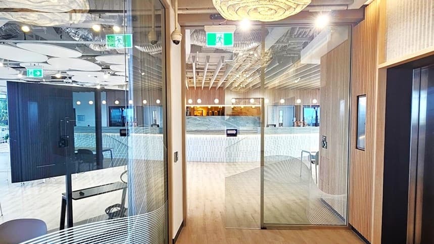 A hallway with a large, white, organic shaped light hanging from the ceiling. On the left is a glass door leading into an office space with tables and chairs and a large screen. On the right is a wall with a set of stainless steel elevator doors. The ceiling is made of light wood panels. The floor is a light brown wood laminate.
