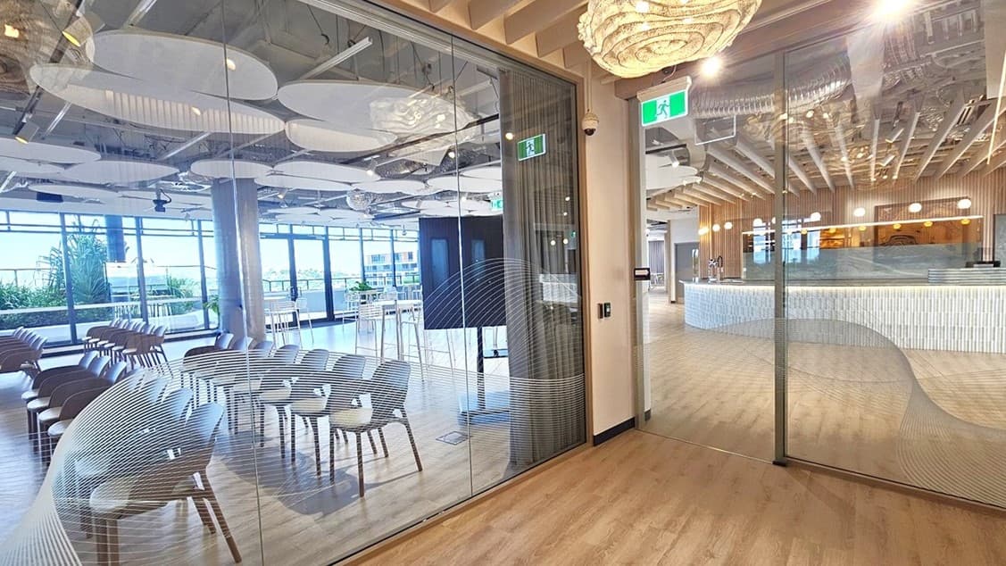 A glass door with a wavy, white pattern on it, leads into a room with a large, white, organic shaped light hanging from the ceiling. The ceiling has a lot of light wood panels. On the left, through the glass door, you can see rows of chairs and a large screen. Behind the chairs is a large window with a view of trees and buildings. To the right of the door, there is another glass door leading into another room with a counter. The floor is wood laminate.