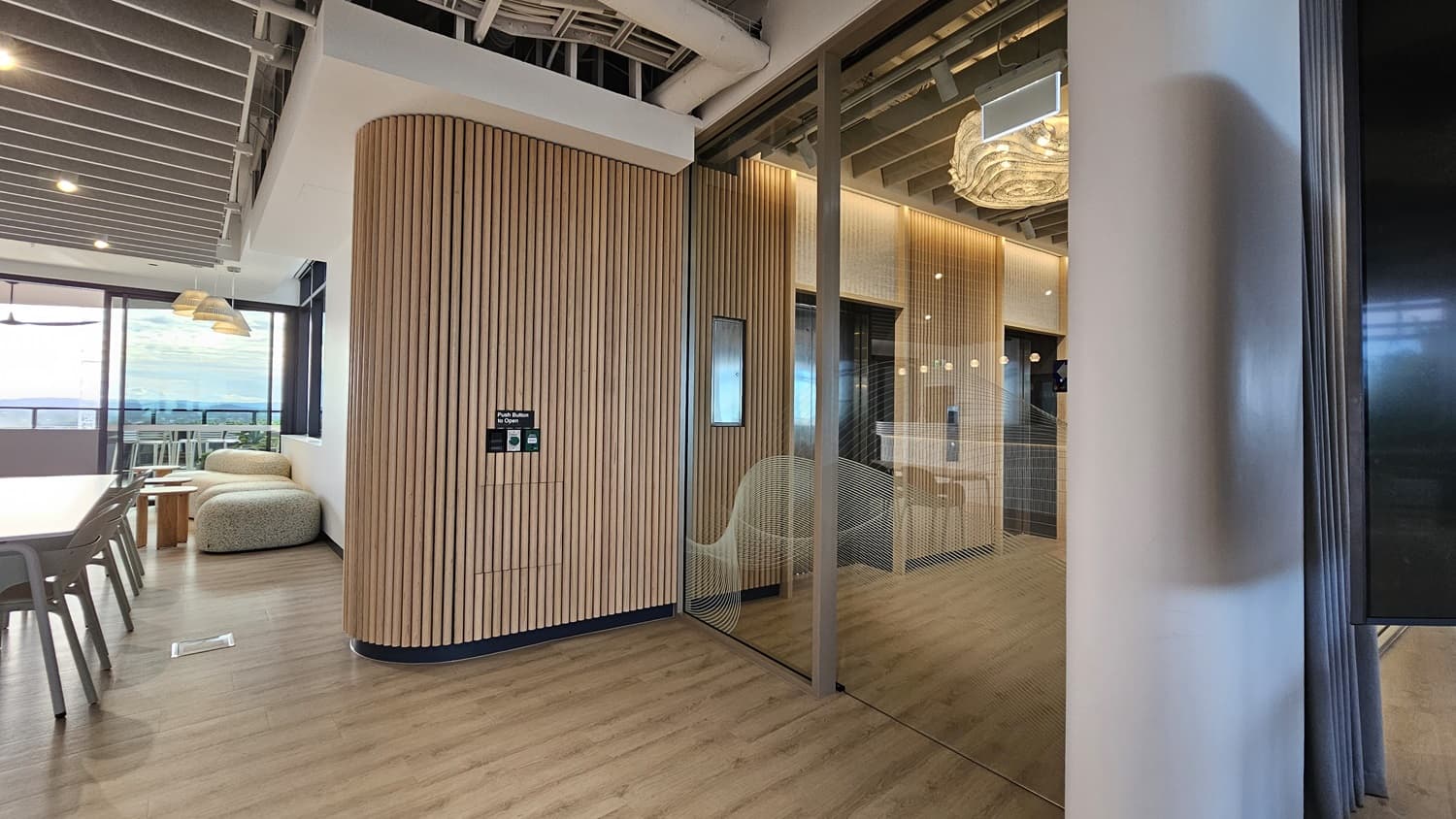 A hallway with a wood-panelled wall on the left, and a glass door on the right leading into a room. The ceiling has wood panels and a large, organic-shaped, light fixture. Through the glass door you can see a large screen, a counter and an elevator. The floor is wood laminate.