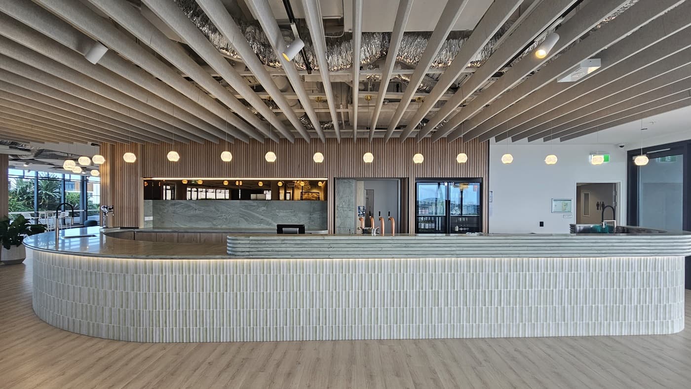 A curved bar with a white tiled front and a grey marble countertop. The bar is lit by a row of round, white pendant lights. The ceiling has wood panels and is lined with rows of spotlights. The floor is wood laminate. On the left is a large window and a small plant in a pot. On the right are two glass doors leading to a room with another counter and a few tables and chairs.