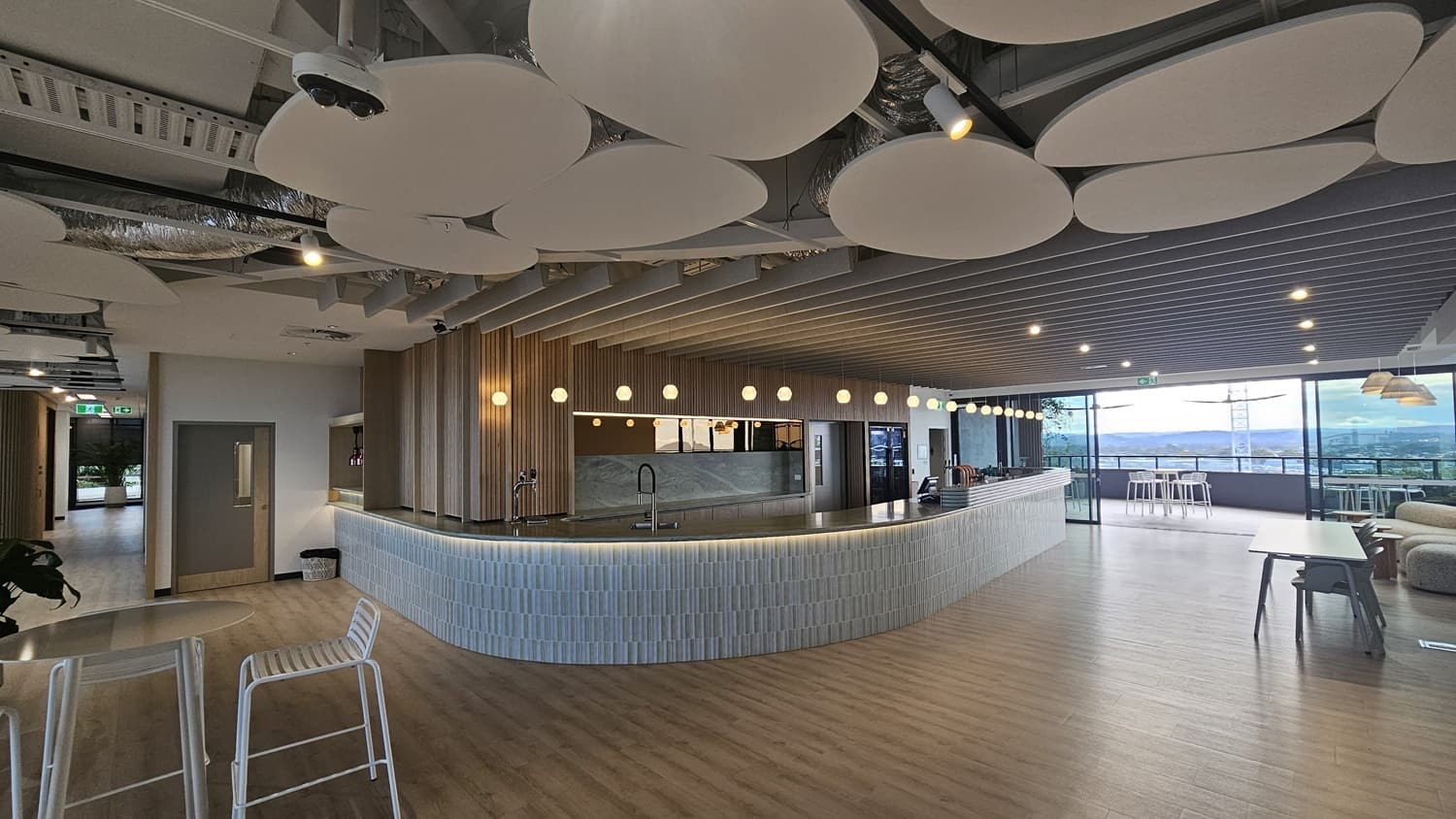 A curved bar with a white tiled front and a grey marble countertop. Behind the bar, there is a wood-panelled wall with a sink and a tap. The ceiling has wood panels and is lined with rows of spotlights. There are also large white, organic shaped lights. The floor is wood laminate. On the left is a doorway, and on the right are glass doors leading to a balcony with a view of trees and buildings.