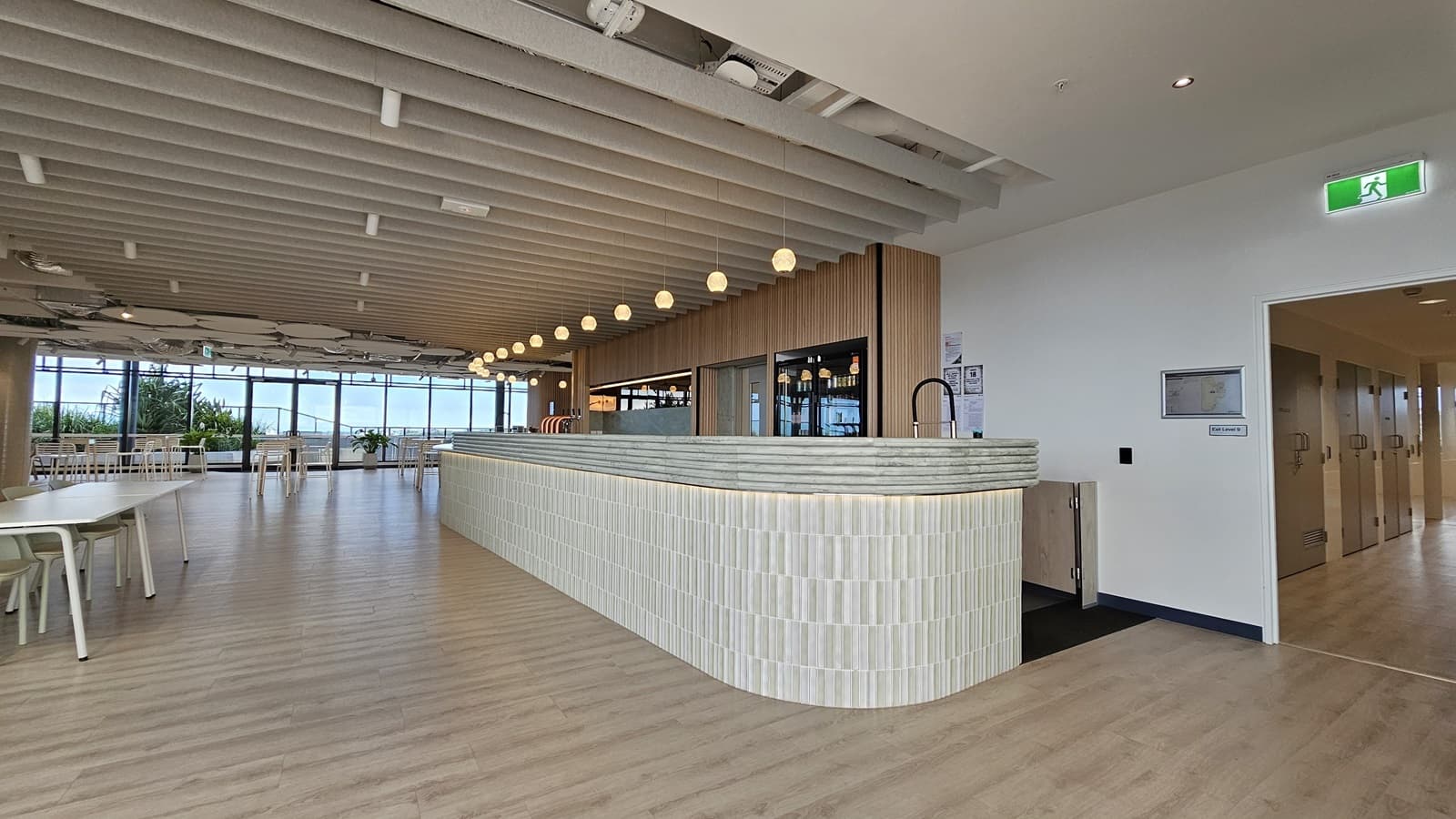 A large, open space with a curved bar in the middle. The bar has white tiles and a grey marble countertop. The ceiling has wood panels and is lined with rows of spotlights. There are also large white, organic shaped lights hanging from the ceiling. To the left of the bar are large windows with views of trees and buildings, and there are tables and chairs for seating. To the right of the bar is a doorway leading to another hallway with a set of doors. The floor is wood laminate.