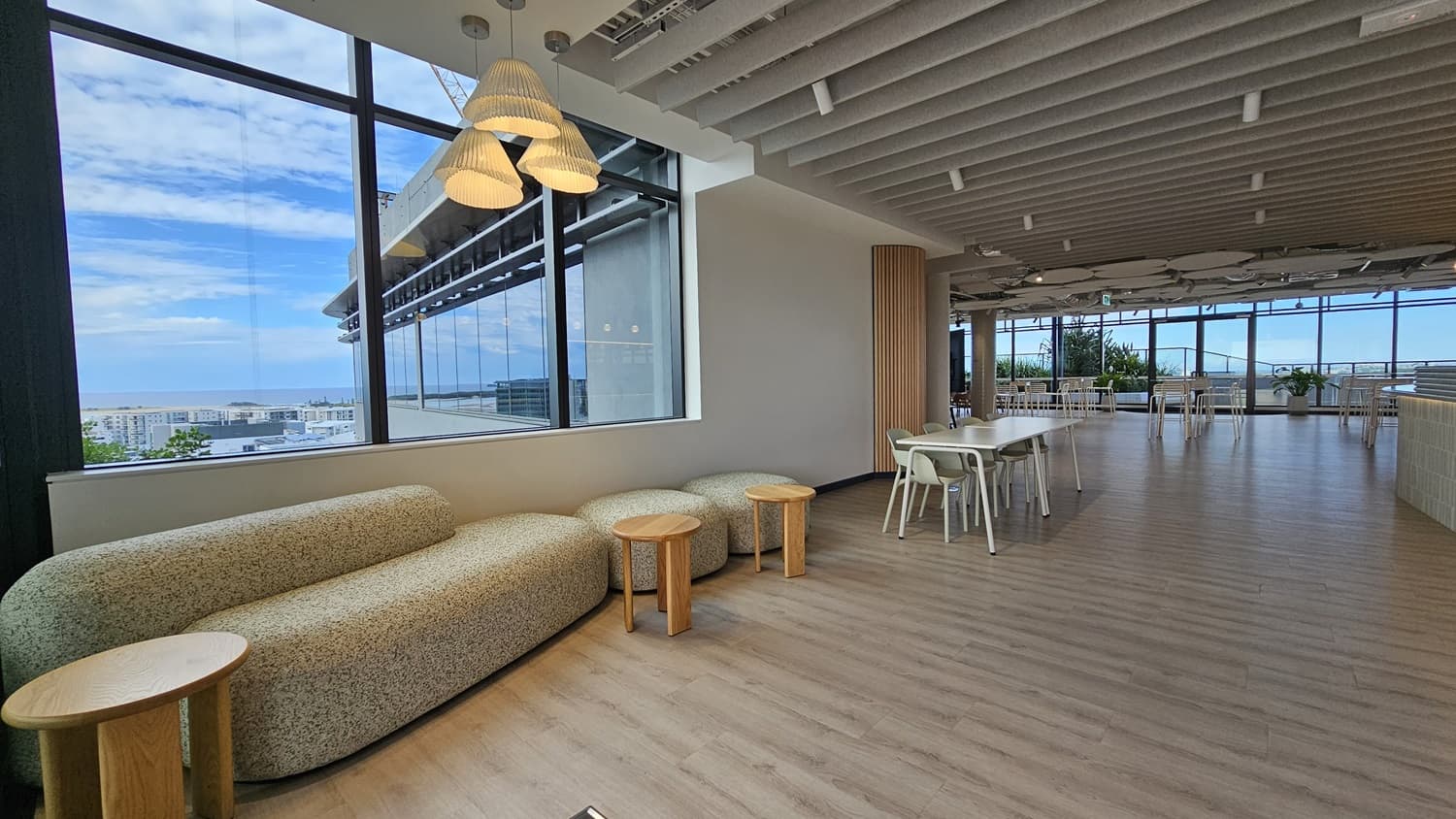 An office lounge area with a large window on the left looking out over a city. There is a large, curved sofa with a small table in front of it. To the right is a long, rectangular table with chairs. The ceiling has wood panels and spotlights. The floor is wood laminate.