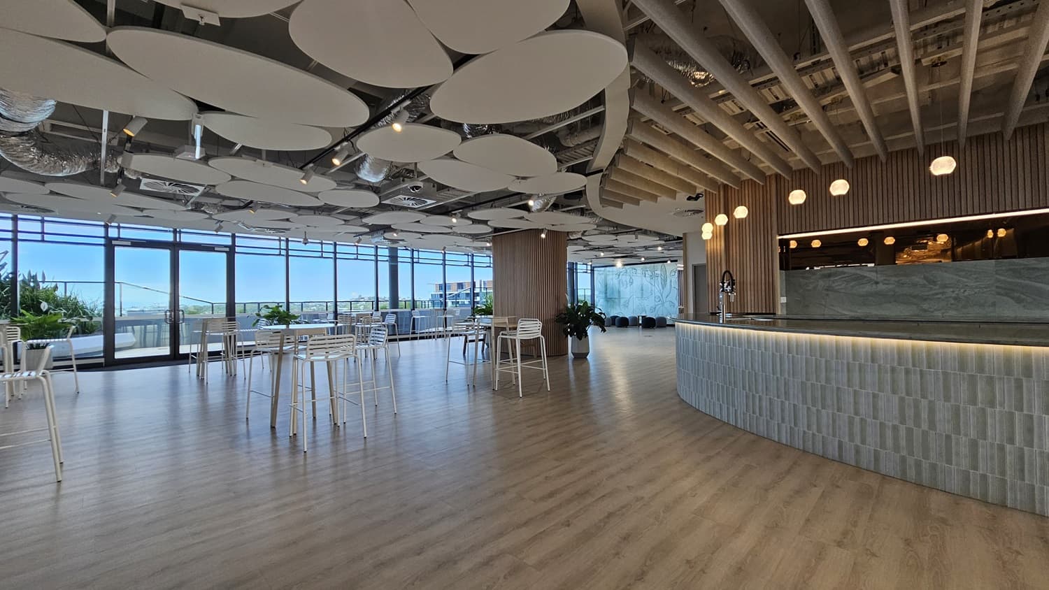 A large, open space with a long, curved bar on the right. The bar has a grey marble countertop and is lined with grey tiles. The ceiling is made of wood panels and has a unique design with many large, circular, white panels. The floor is wood laminate. On the left is a large window with a view of a city. There are several tables and chairs in the middle of the room, and there are also some small plants.
